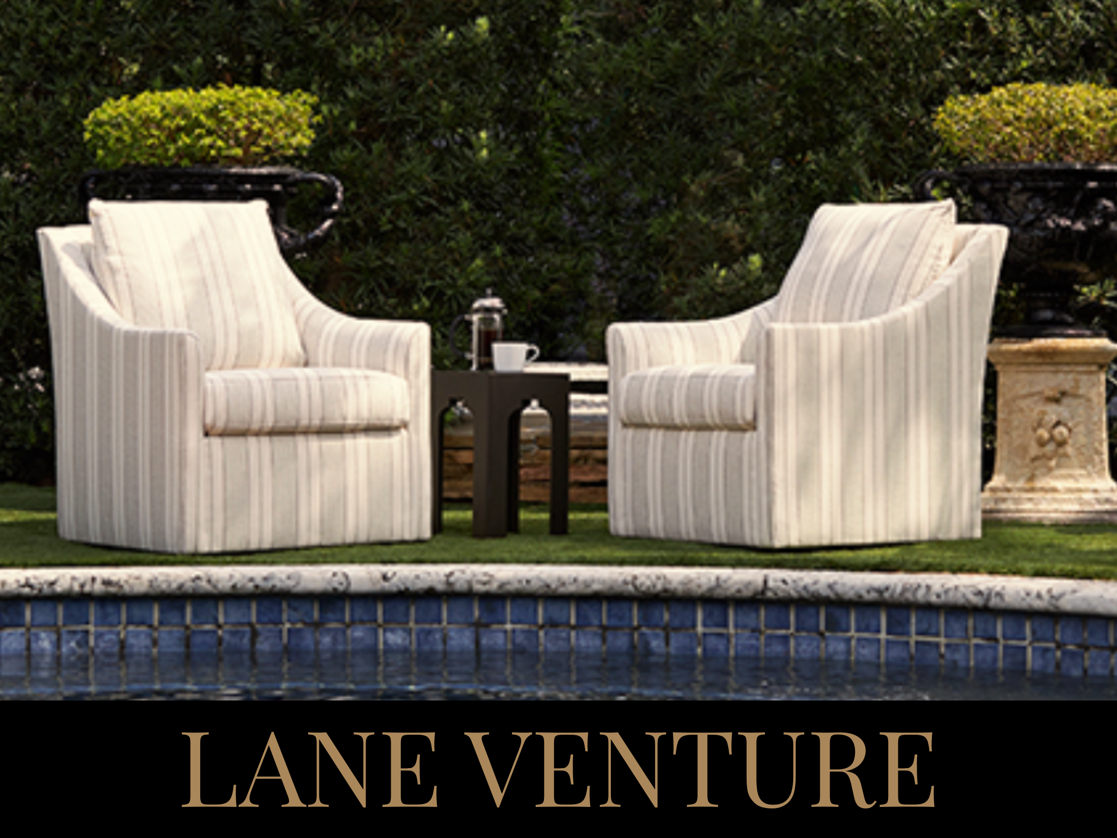 Two armchairs with striped upholstery facing each other with a small black side table between them, outdoor pool in the foreground, lush greenery in the background, and "LANE VENTURE" text at the bottom.