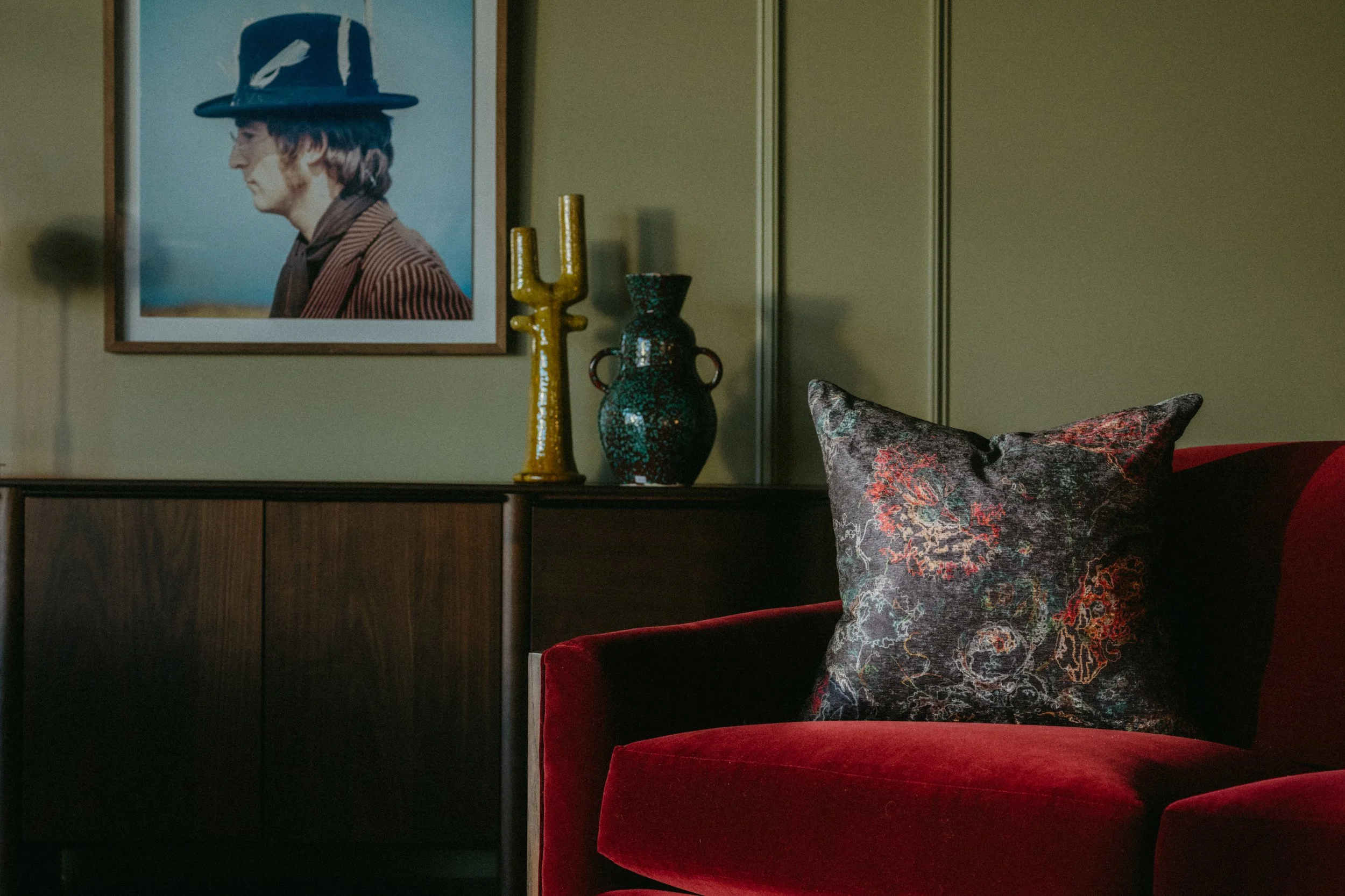 A framed portrait of John Lennon sits above a curated red velvet armchair with a patterned throw pillow at The Home Studio Collective showroom. 