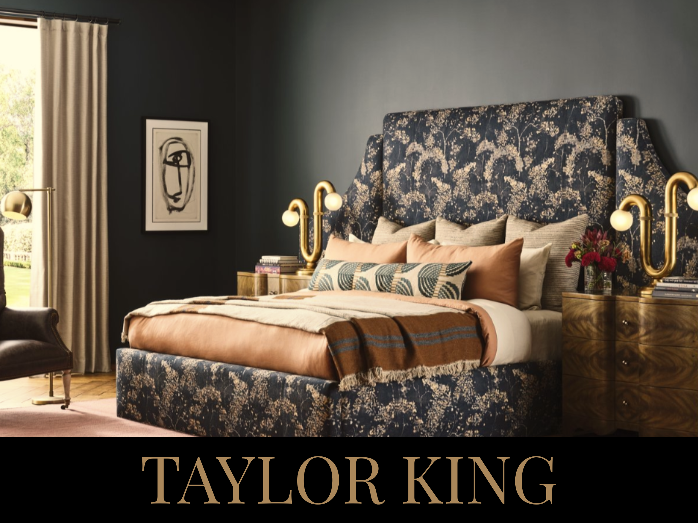 A bedroom with a dark blue floral upholstered bed, gold bedside tables, and golden wall-mounted lamps with white globes. The bed has various pillows and a brown and orange throw blanket. There is a beige curtain over a window and a framed abstract bl