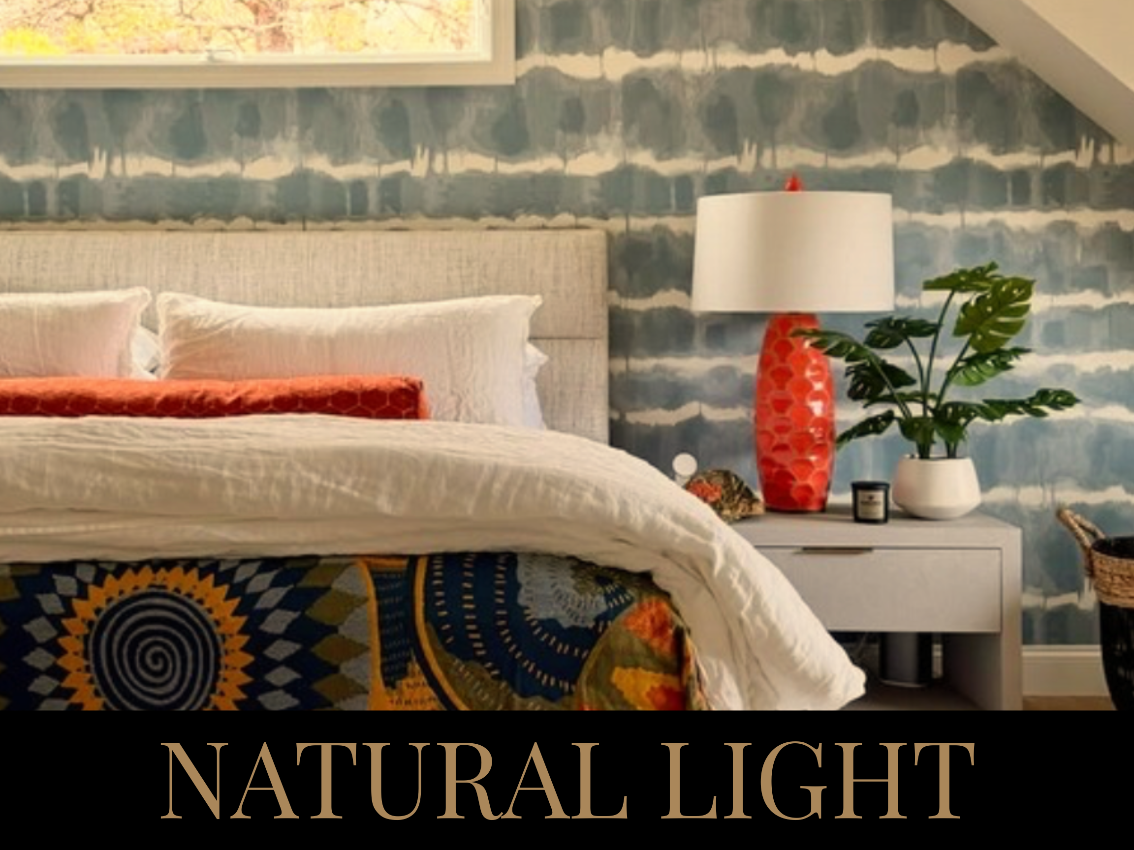 A stylish bedroom with a bed, patterned blanket, white pillows, a nightstand with a lamp, potted plant, and decorative items, featuring a blue and white painted wall background and natural light coming through a window.