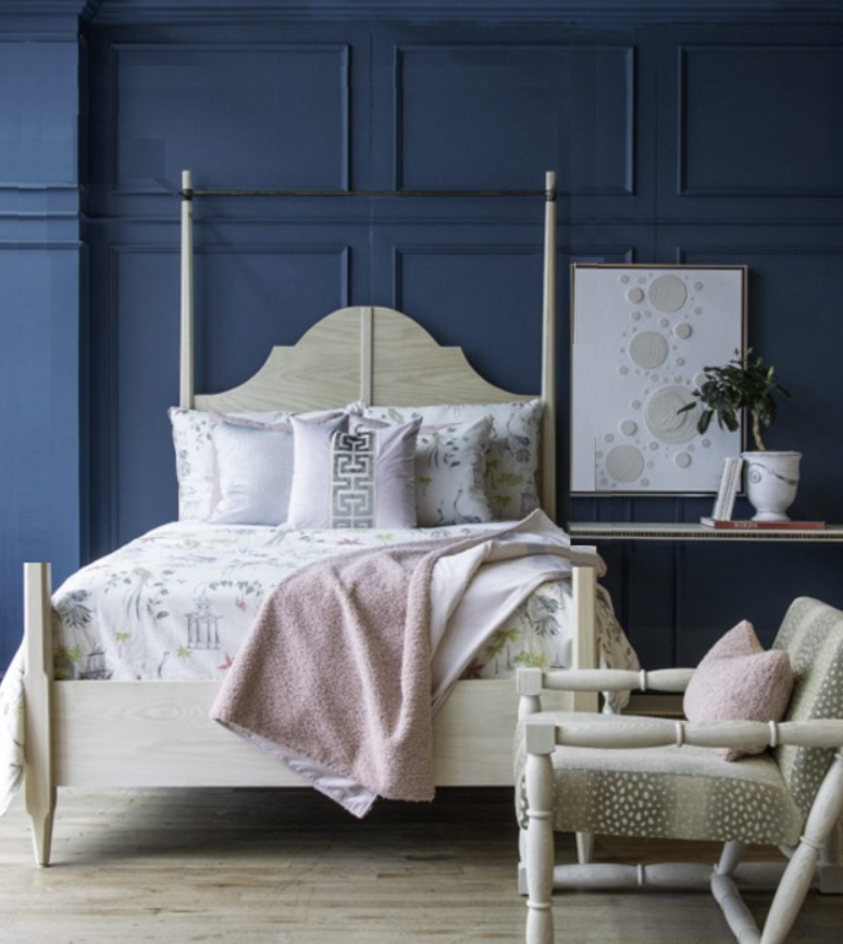 A bedroom with a navy blue paneled wall, a light wood bed frame with bedding, decorative pillows, and a pink throw blanket. There is a matching armchair with a pink pillow, and a side table with a potted plant and artwork.