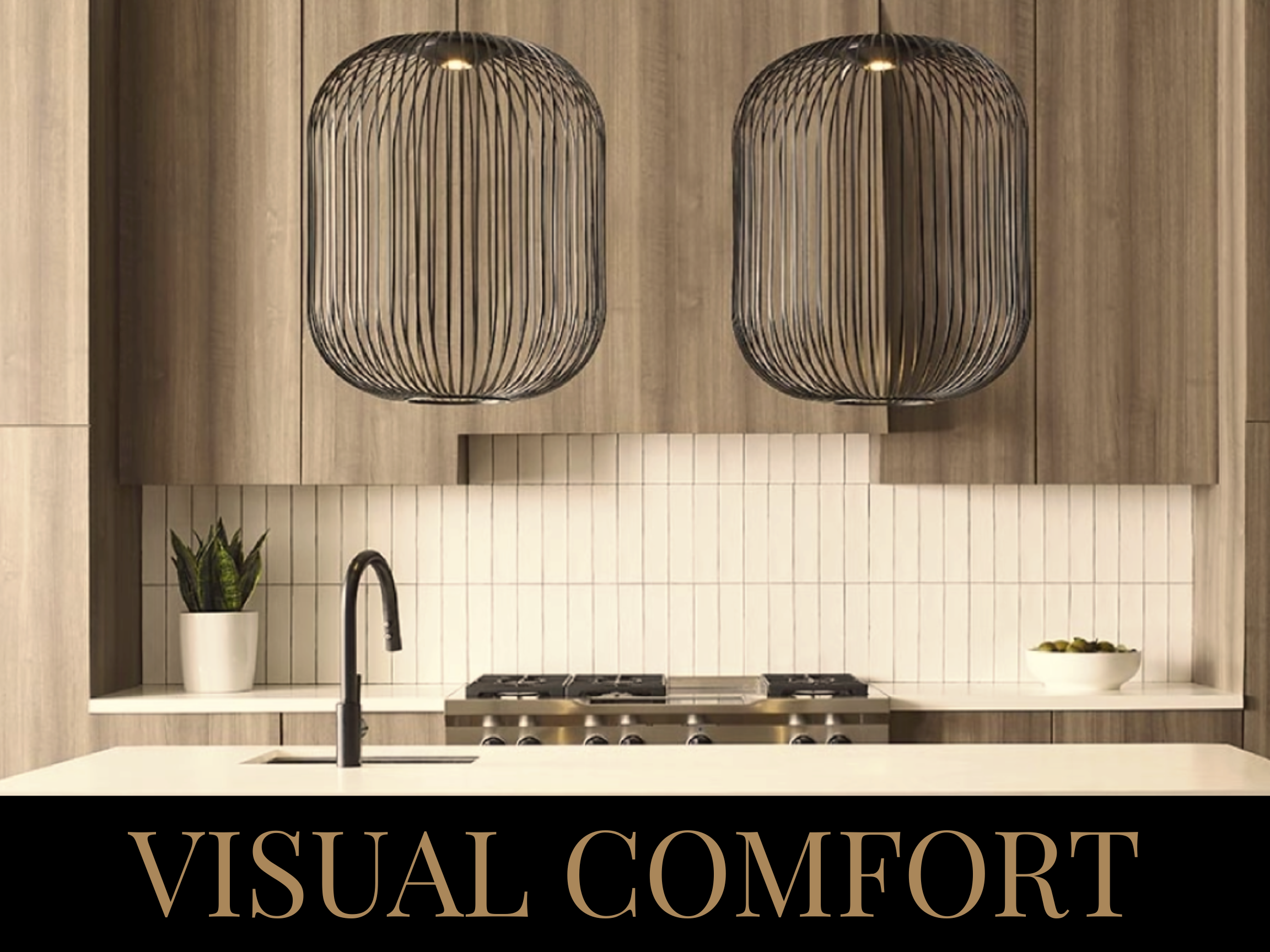 Modern kitchen with two large black pendant lights, light wood cabinets, a white countertop with a sink, potted plant, and a bowl of fruit, with the words 'VISUAL COMFORT' at the bottom.