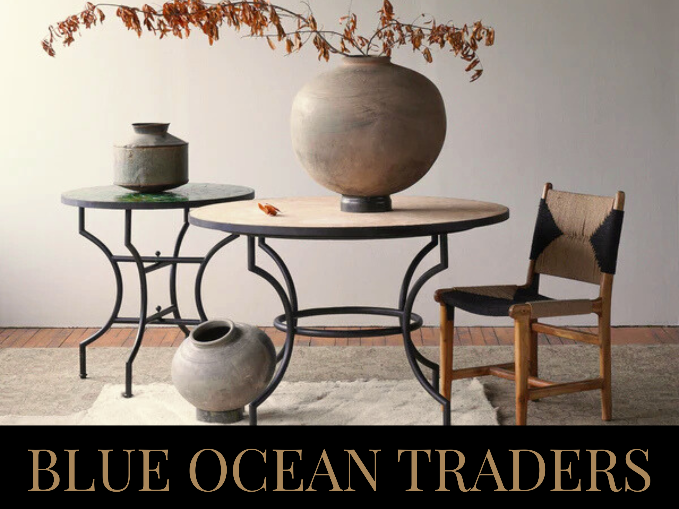 Interior decor with two round tables, one with a large gray ceramic vase with dried branches, and the other with a small piece of decor on top. A wooden chair with black and beige upholstery is beside the tables. The background is a plain wall, and t