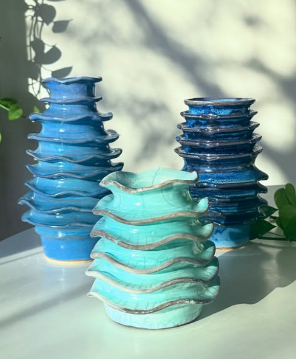 Three stacks of glazed ceramic bowls in shades of blue and turquoise with irregular wavy rims, set against a light background with shadows and greenery.