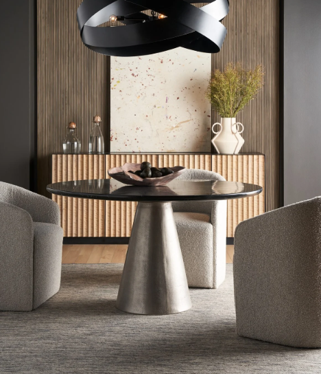 Modern living room with a round black dining table, two gray armchairs, a wood-paneled wall, a large abstract painting, a white vase with greenery, and decorative bottles, under a black sculptural pendant light.