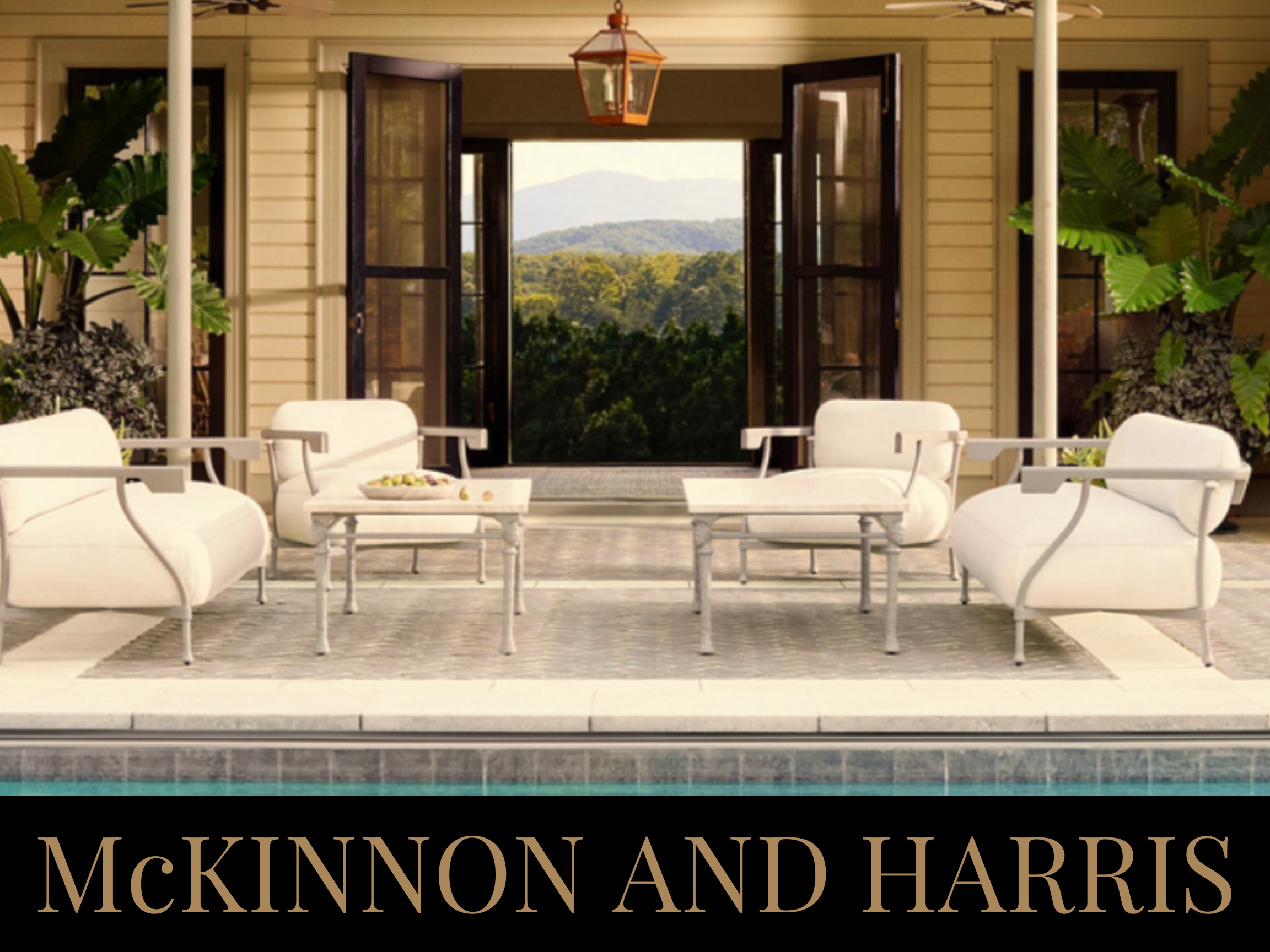 Outdoor patio with white cushioned furniture facing a house with open doors revealing a view of trees and mountains in the background.