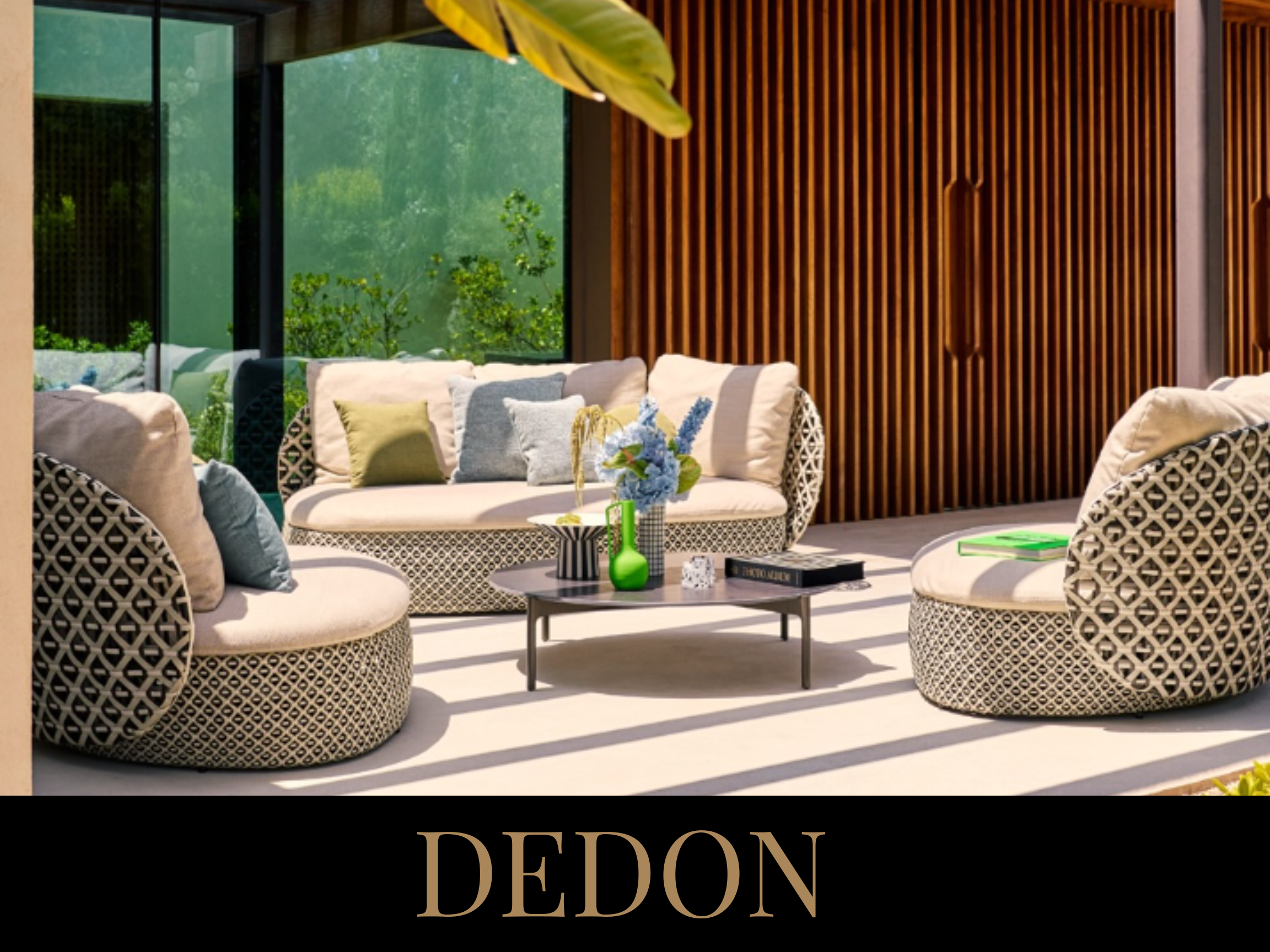 Modern outdoor patio with patterned lounge chairs and sofa, a black coffee table with vases and books, and a wooden slat wall, with large sliding glass doors showing greenery outside.