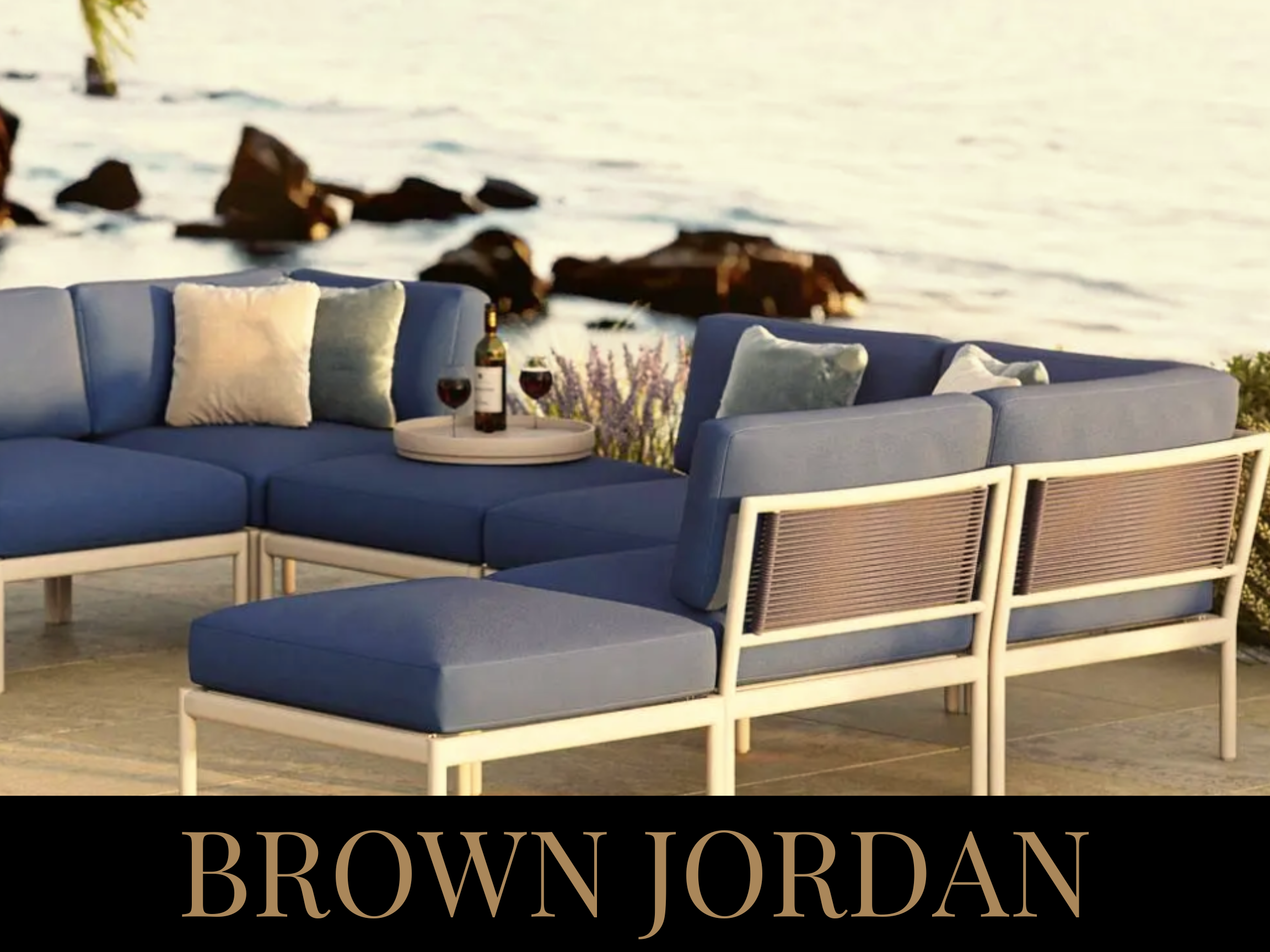 Outdoor patio furniture with blue cushions and pillows, set beside a body of water with rocks, during sunset or sunrise. The furniture includes a sectional sofa and an ottoman, with a tray holding wine bottles and glasses on the sofa.