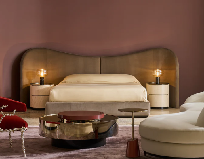 Modern bedroom with a large beige headboard, two bedside tables with lamps, a round coffee table, and a white lounge chair.