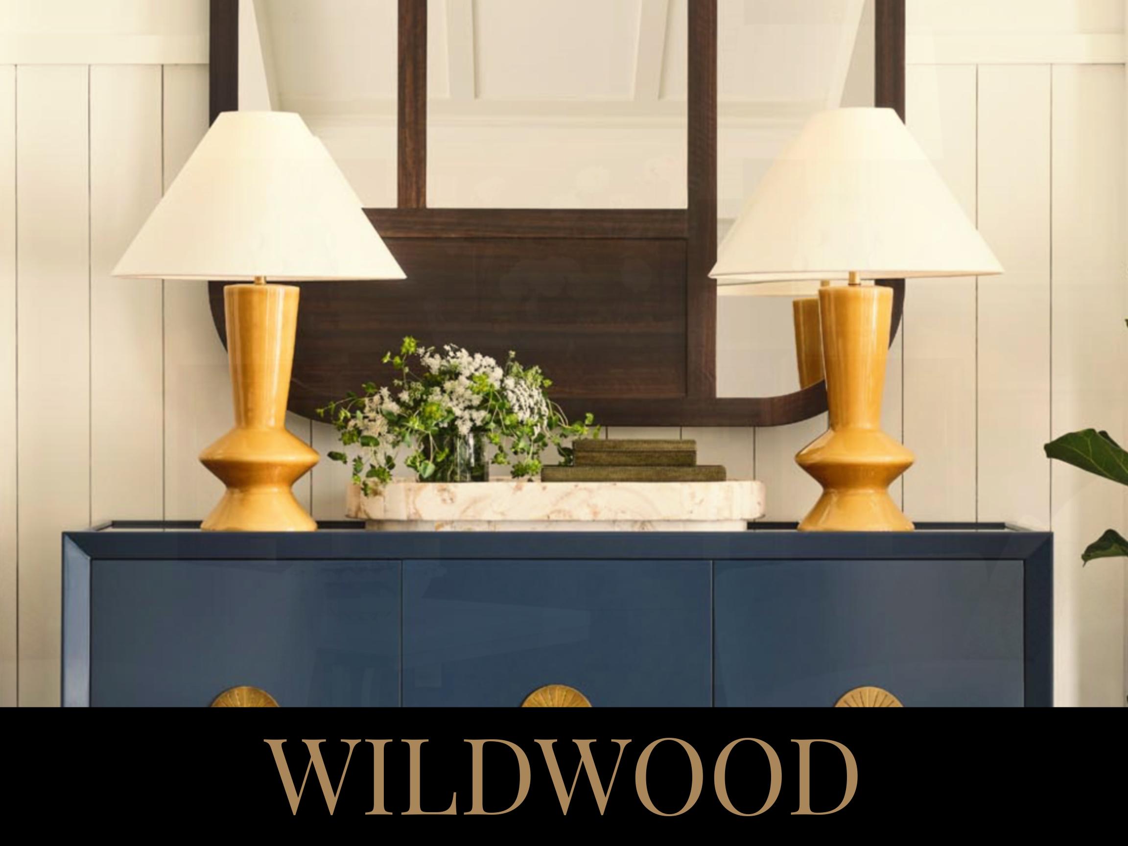 A blue sideboard with two yellow lamps, a white marble tray with green foliage, and a few books. Behind is a large wooden mirror on a white paneled wall. The word 'WILDWOOD' is written at the bottom.