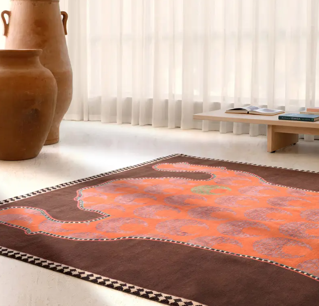 A living room with a patterned orange and pink rug, large earthenware vases, sheer white curtains, and a low wooden table with books and an open magazine.