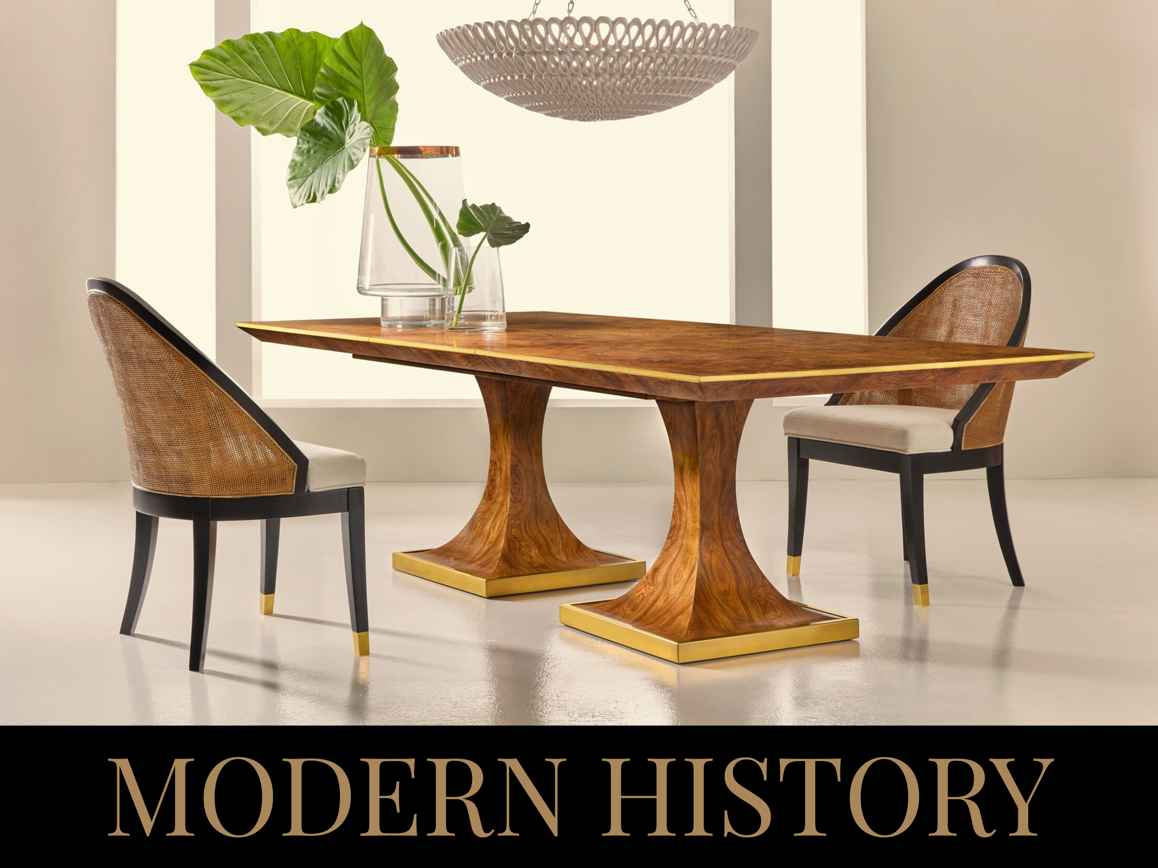 A wooden dining table with a curved base, two chairs with woven backs, and a glass vase with large green leaves, in a room with natural light and a minimalist decor.