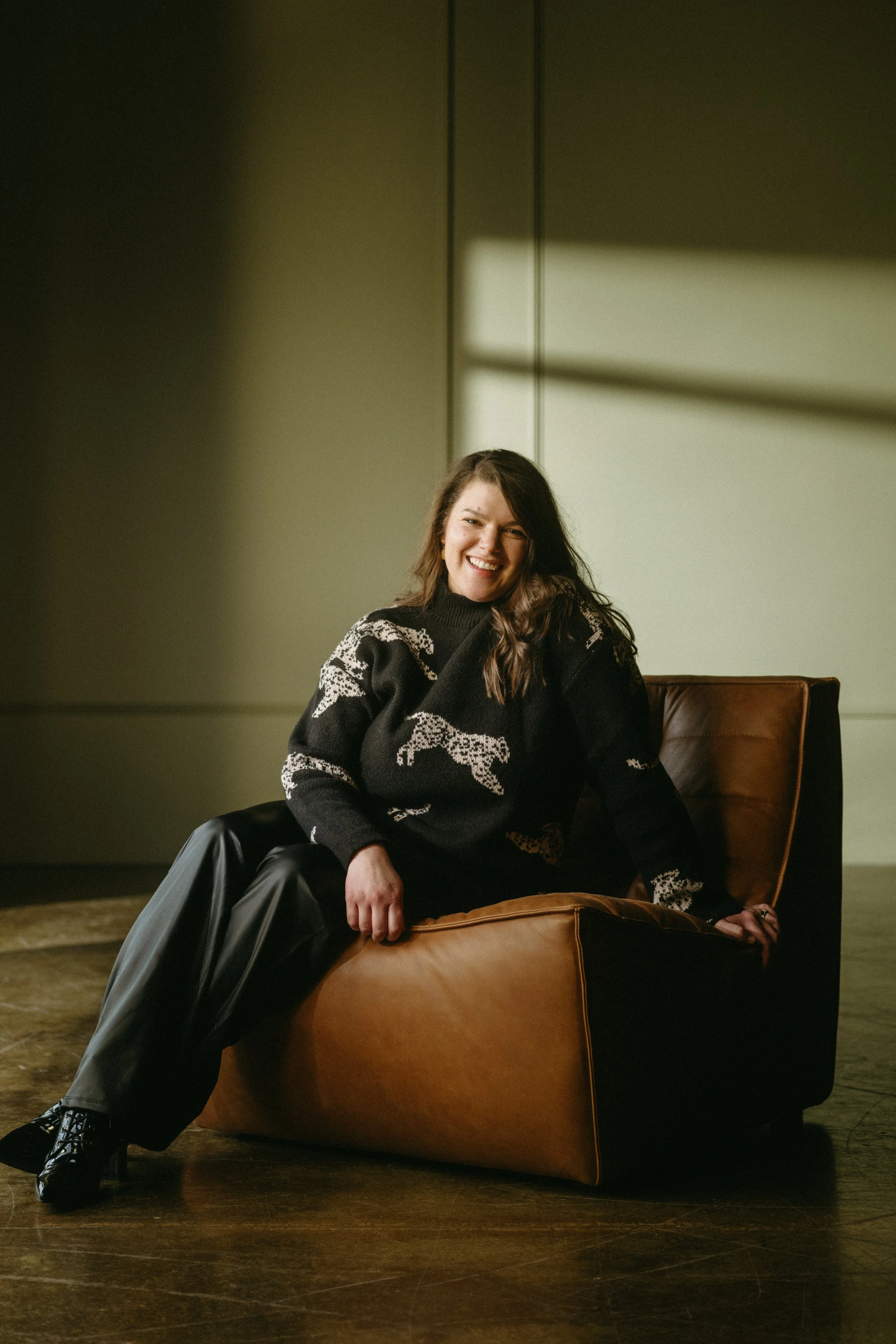 A woman sitting on a brown leather chair, smiling, in a dimly lit room with a window casting shadows on the wall.