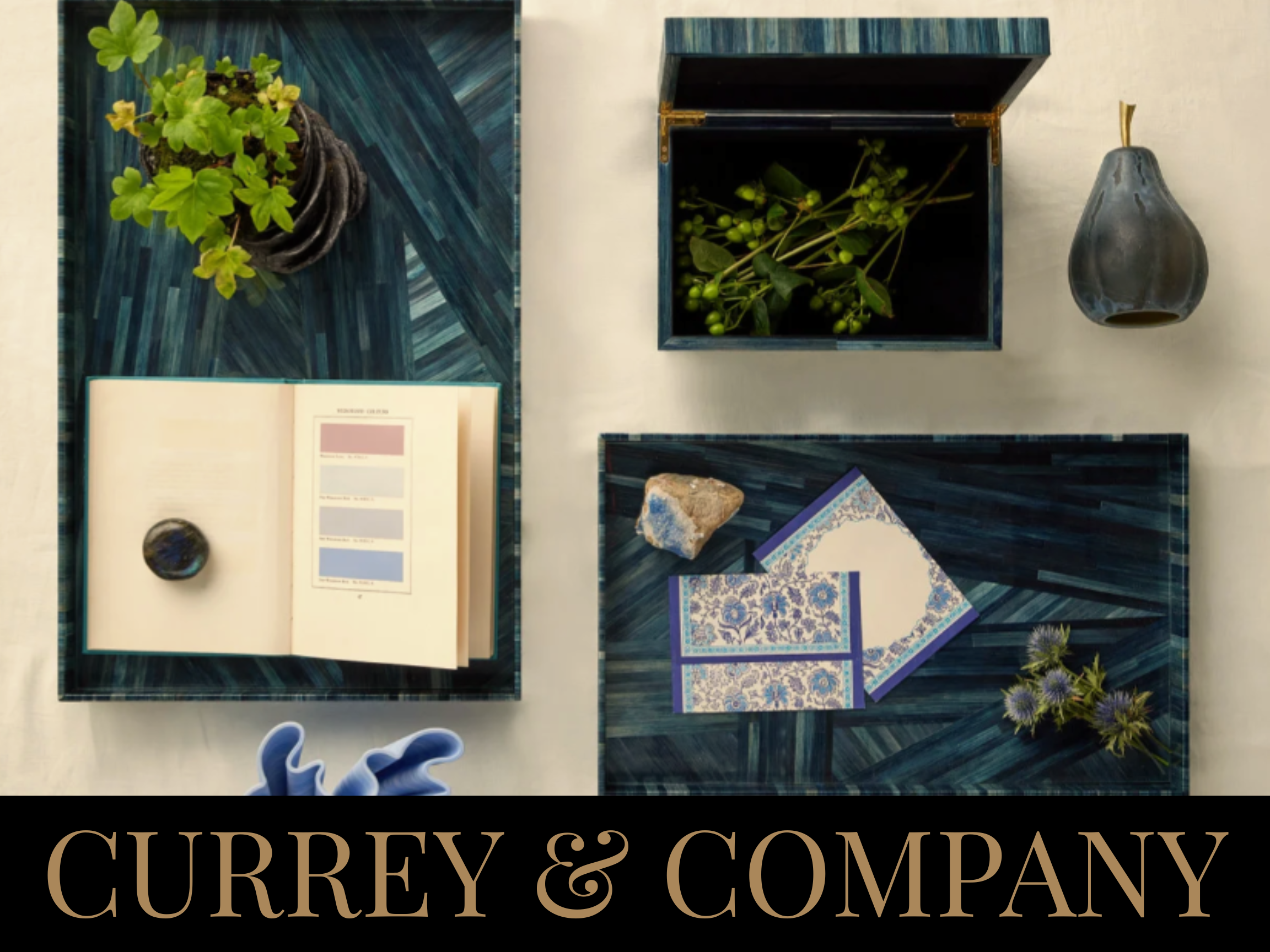 A flat lay of decorative items on two blue geometric-patterned trays. The items include a small potted plant, an open book with a marble-like paperweight, some patterned stationery, a small stone, and some thistle flowers. The background features a l