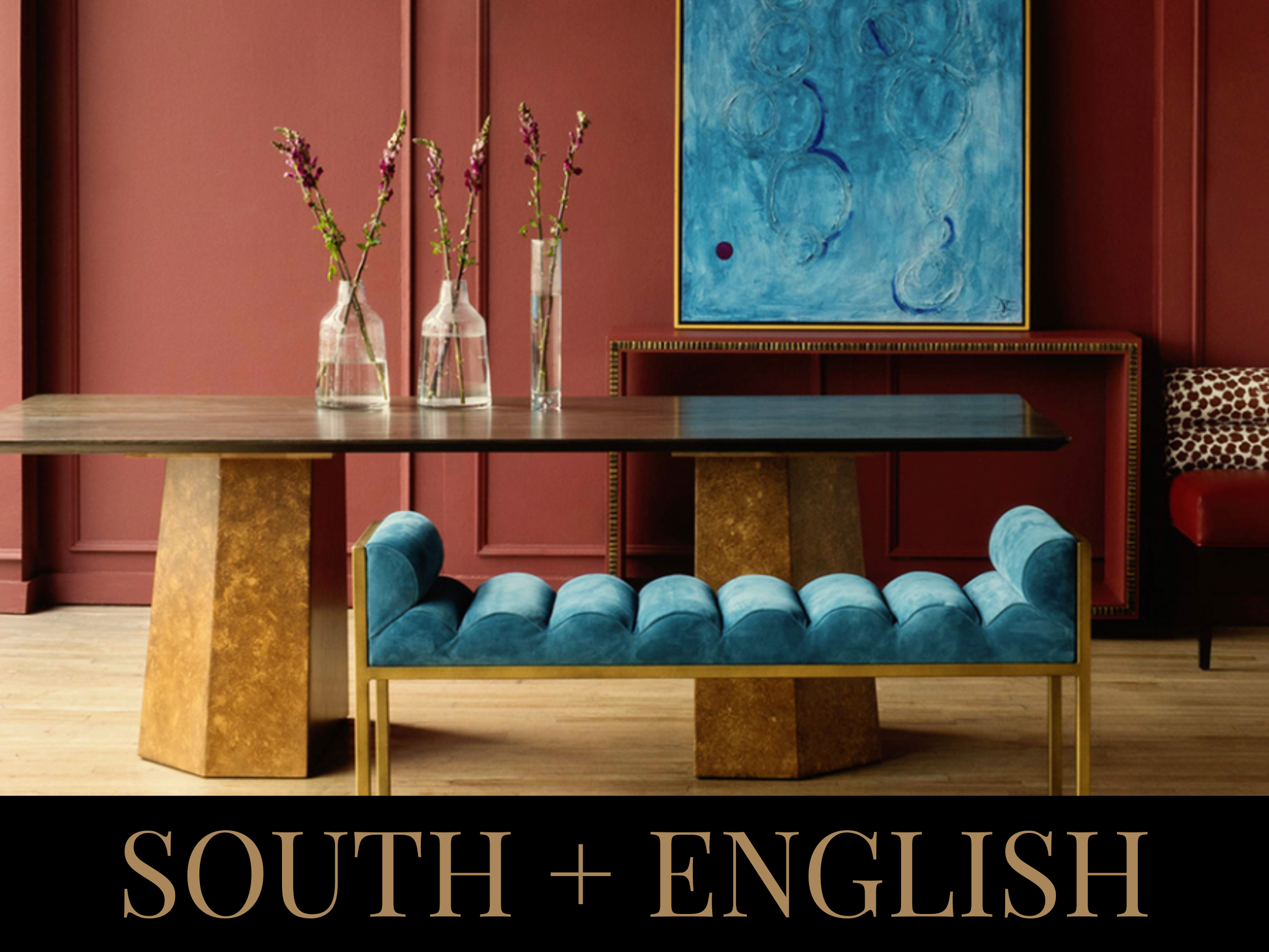 Interior of a room with red paneled walls, a wooden table with three vases of flowers, a framed blue abstract painting, a gold and blue upholstered bench, and a red sofa with patterned cushion. Text reads "SOUTH + ENGLISH" at the bottom.