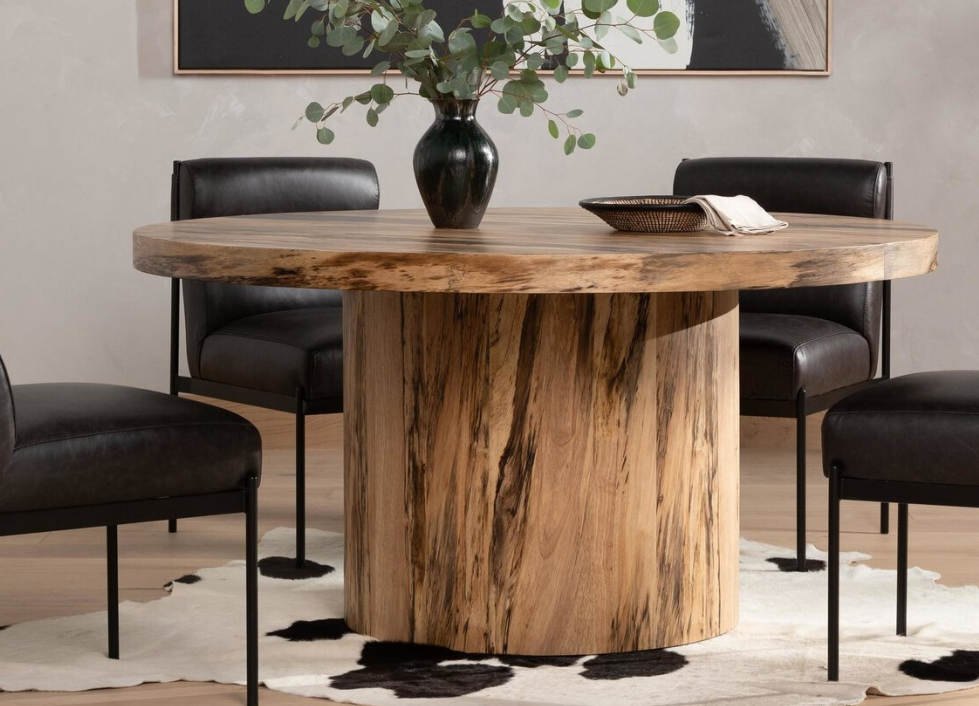 A round wooden dining table with a cylindrical base, surrounded by four black leather chairs, on a black and white cowhide rug, with a black vase filled with green eucalyptus leaves on top, in a modern dining room.