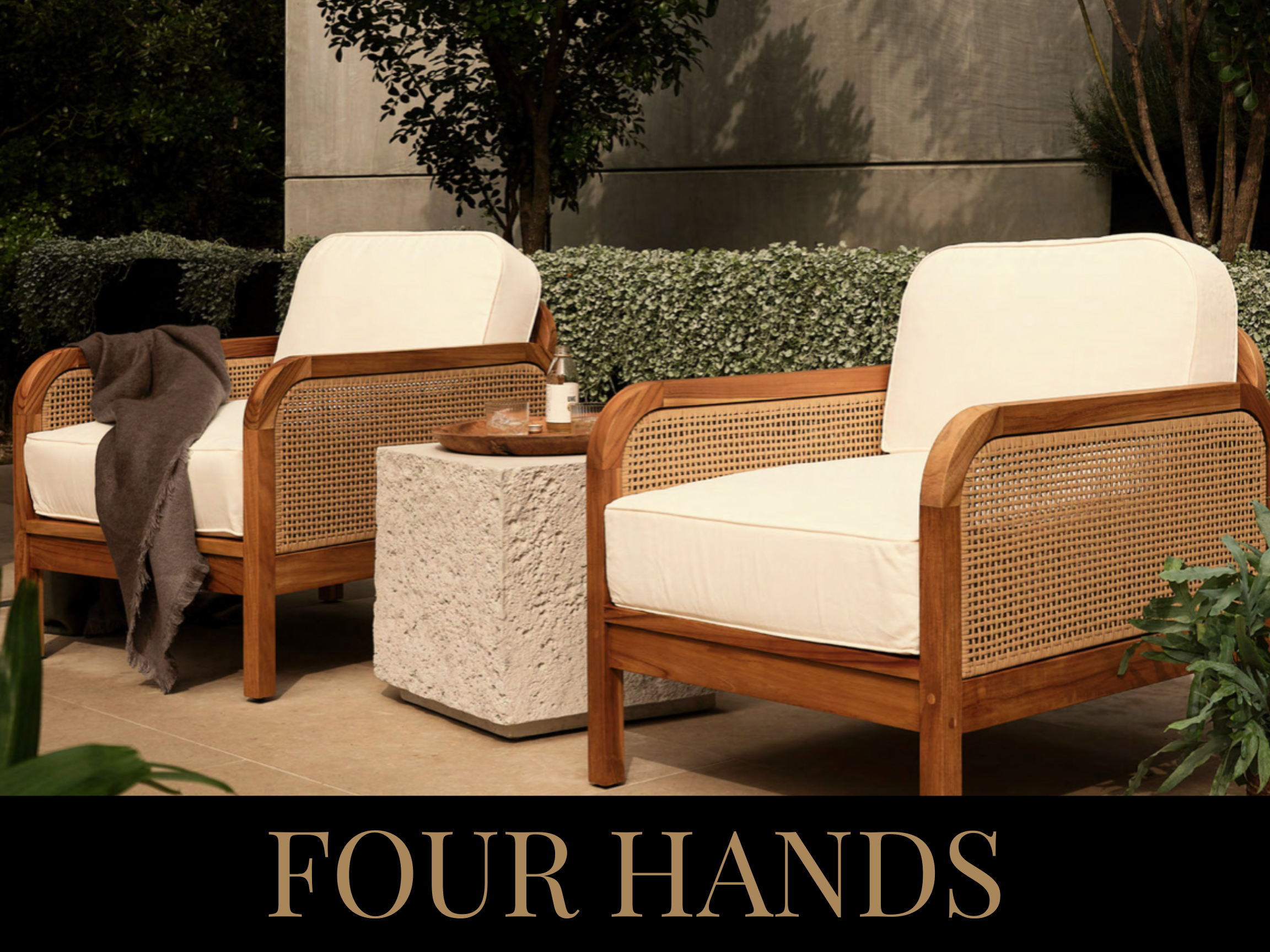 Two white cushioned armchairs with wooden frames and cane sides are placed outside near a stone table with a tray, bottles, and glasses on it, against a background of green bushes and a wall.