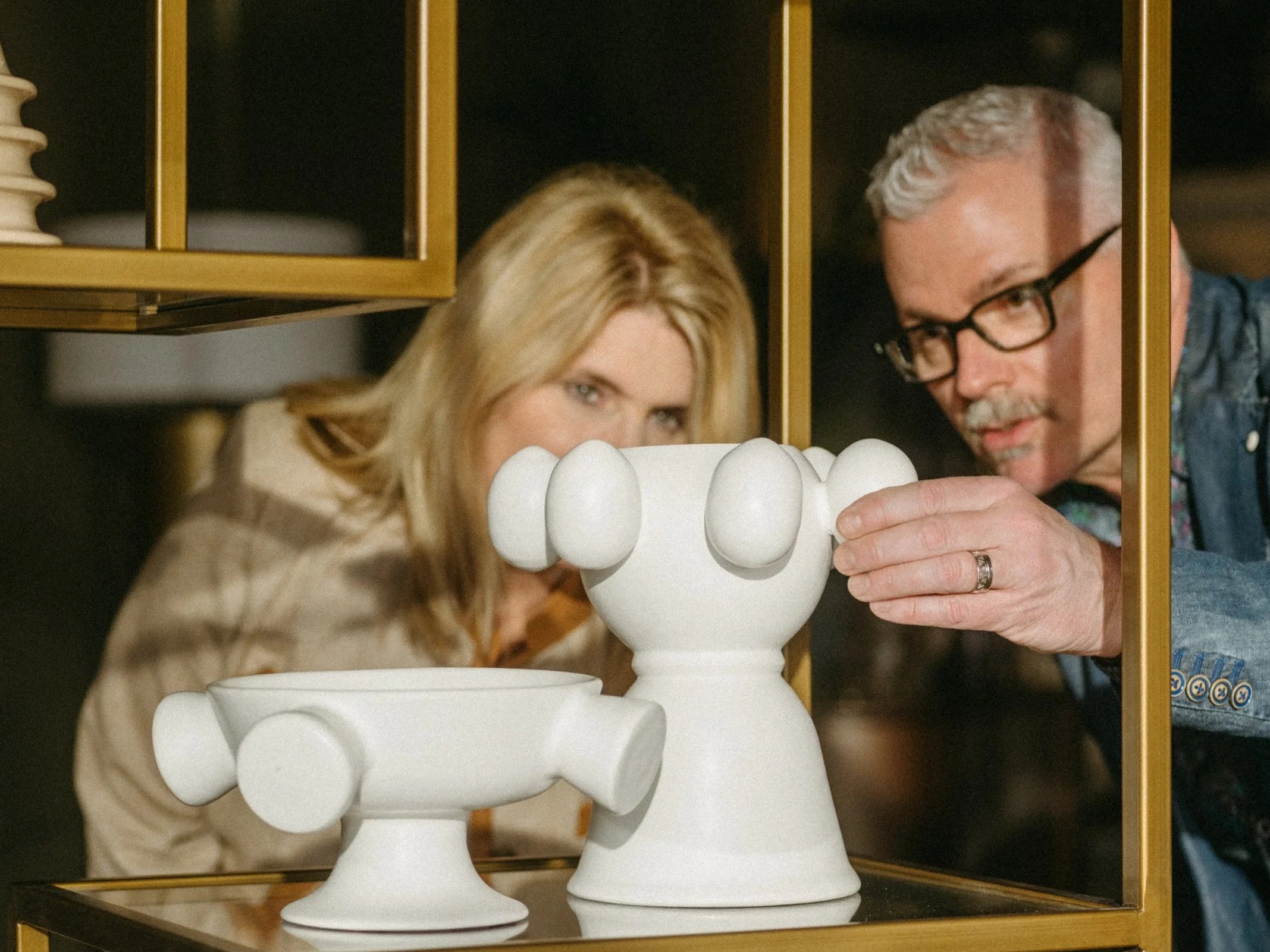 A man and woman observe two white ceramic sculptural objects on a glass shelf, with golden framing. The objects resemble stylized animals or abstract forms.