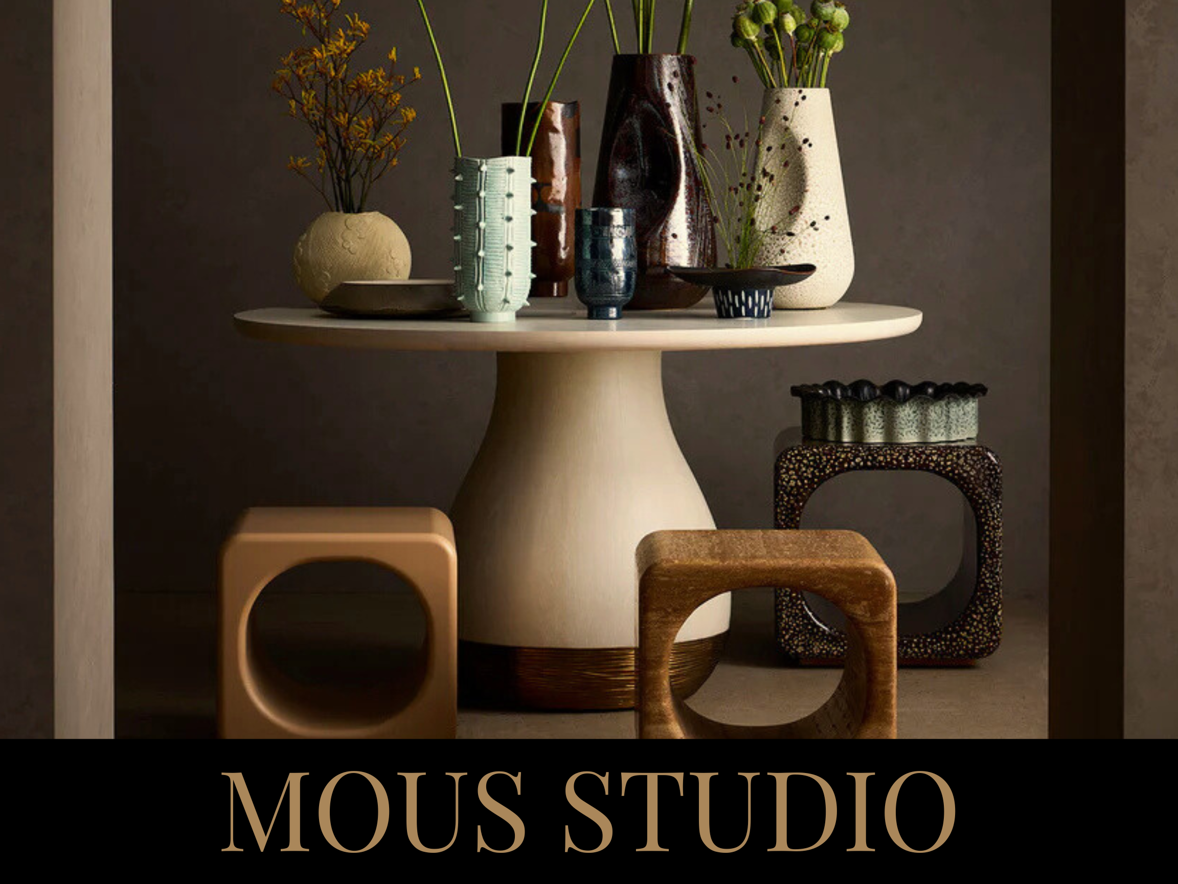 Decorative pottery vases and bowls on a beige round table with black text 'MOUS STUDIO' below. The vases contain various plants with green and brown foliage, set against a dark textured background.