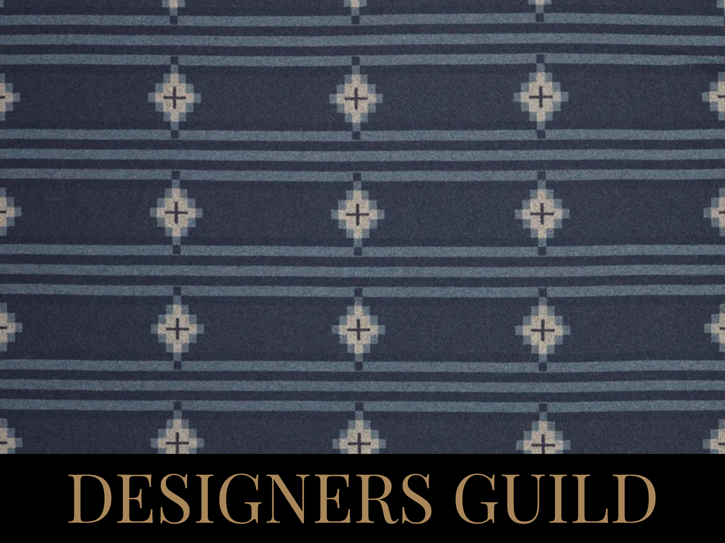 A blue patterned fabric with white geometric designs. The fabric has horizontal stripes with diamond shapes interspersed. The words "DESIGNERS GUILD" are written in large, beige letters at the bottom of the image.