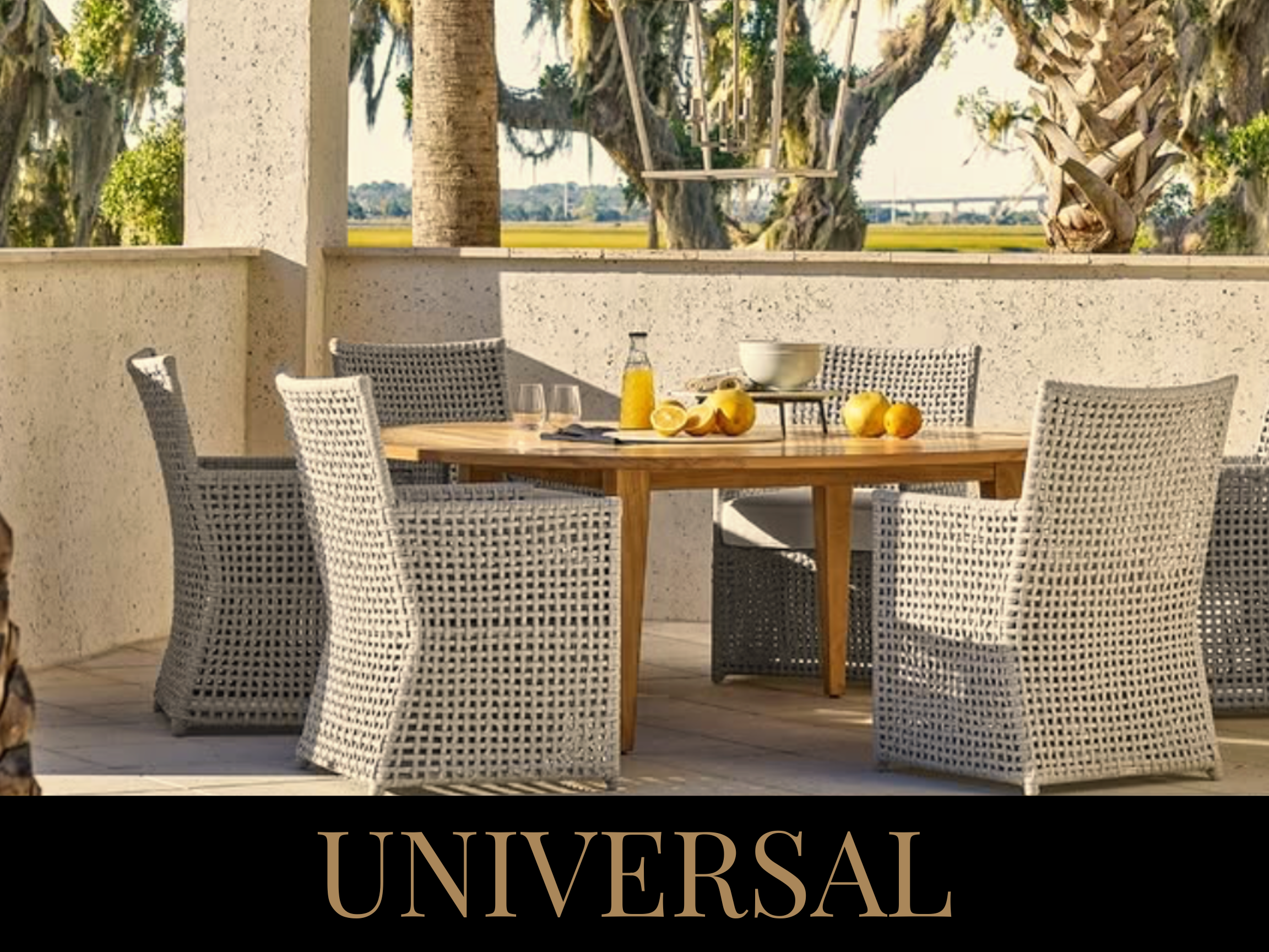 Outdoor patio with a round wooden table set with a bowl, bottle, glasses, and lemons, surrounded by six wicker chairs, with a view of trees and open fields beyond a stucco wall.