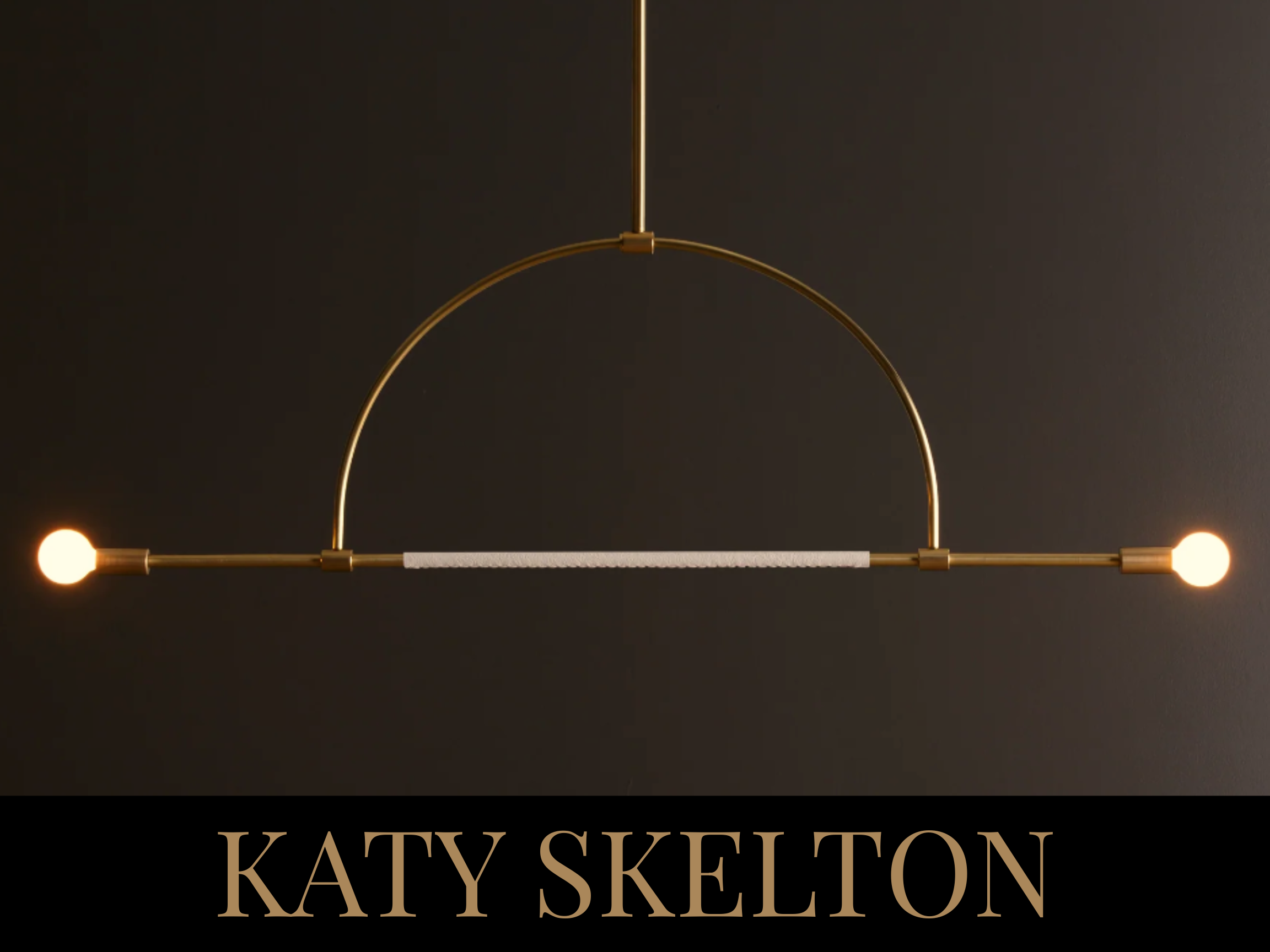 A modern hanging light fixture with two bright bulbs and gold accents, mounted on a dark wall, with the text 'KATY SKELTON' at the bottom.