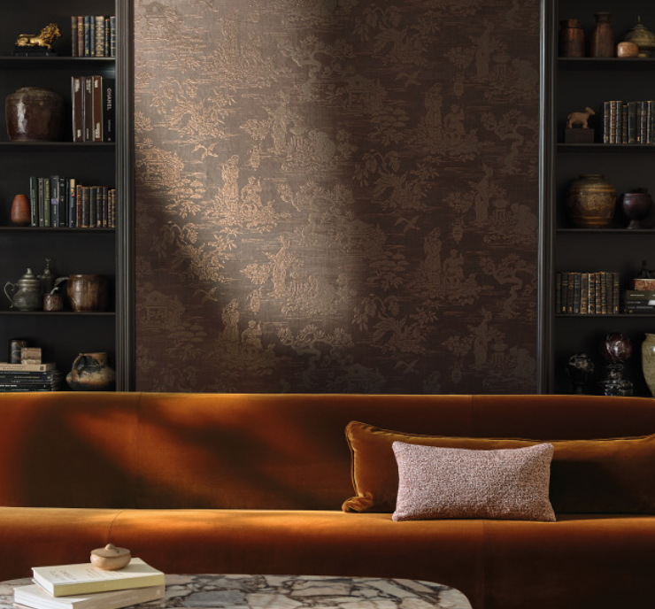 A living room with a brown velvet sofa, a grey pillow, a marble-top table with books and a small decorative bowl, dark wooden wall shelves filled with books and decorative objects, and textured brown wallpaper.