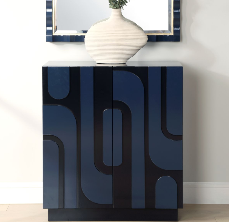 A dark blue modern cabinet with an abstract black pattern, topped with a large white ceramic vase holding green foliage, and a rectangular mirror above it on a light-colored wall.
