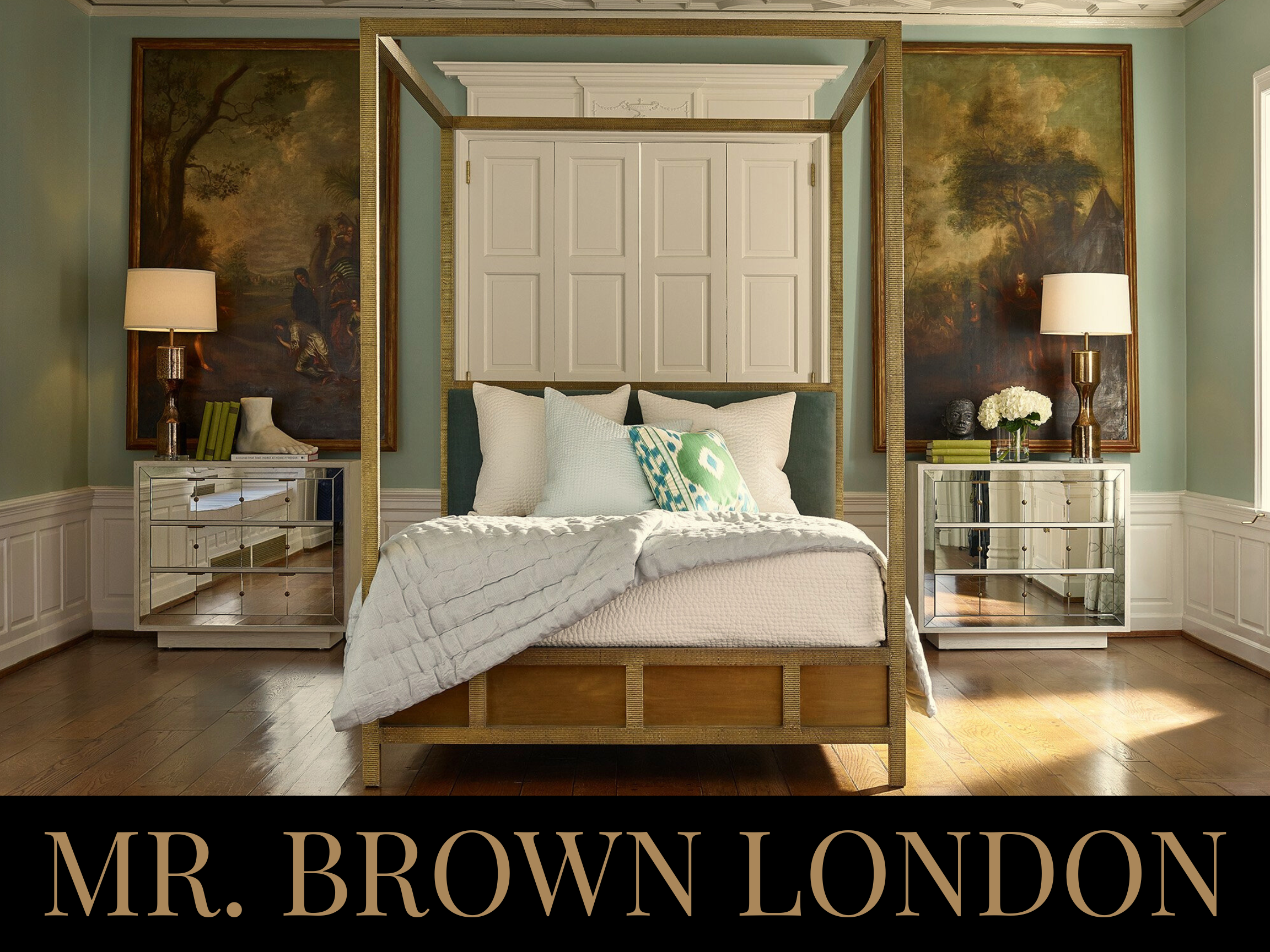 A luxurious bedroom with a four-poster bed, white bedding, decorative pillows, two mirrored nightstands with lamps, paintings on the walls, and wood flooring.