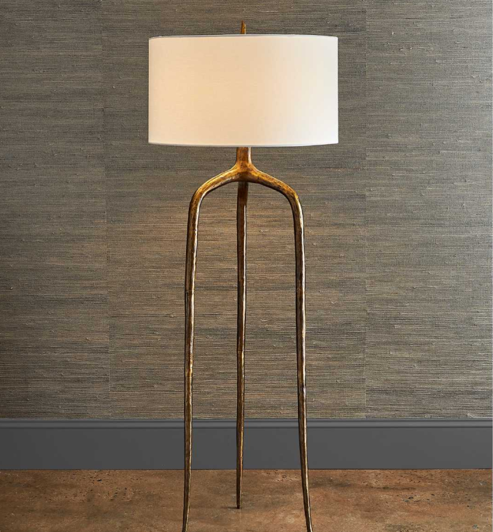 A floor lamp with a wooden tripod base and a white lampshade, standing on a wooden floor against a textured gray wall.