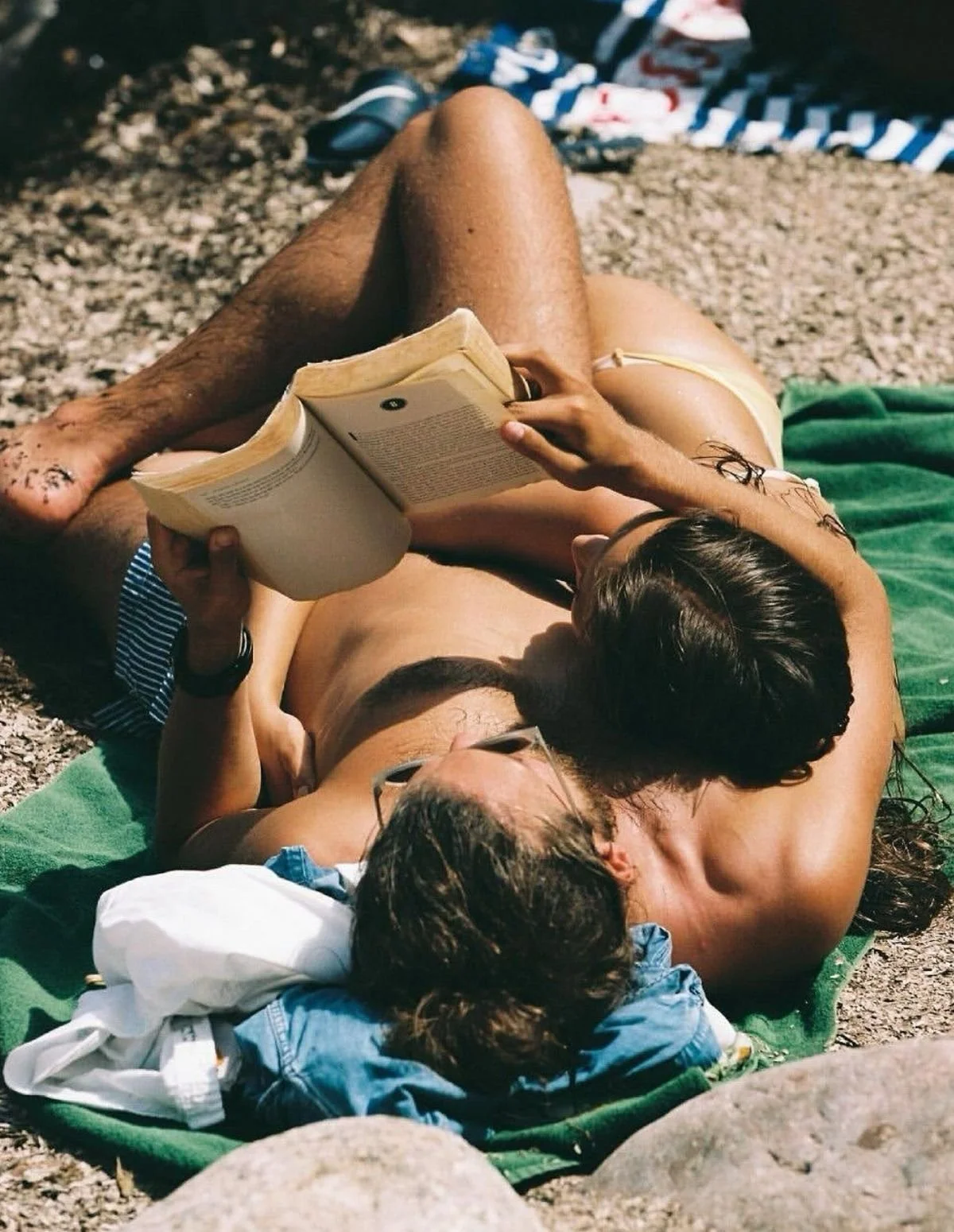 Two people are lying on a green towel on a rocky beach, reading a book together.