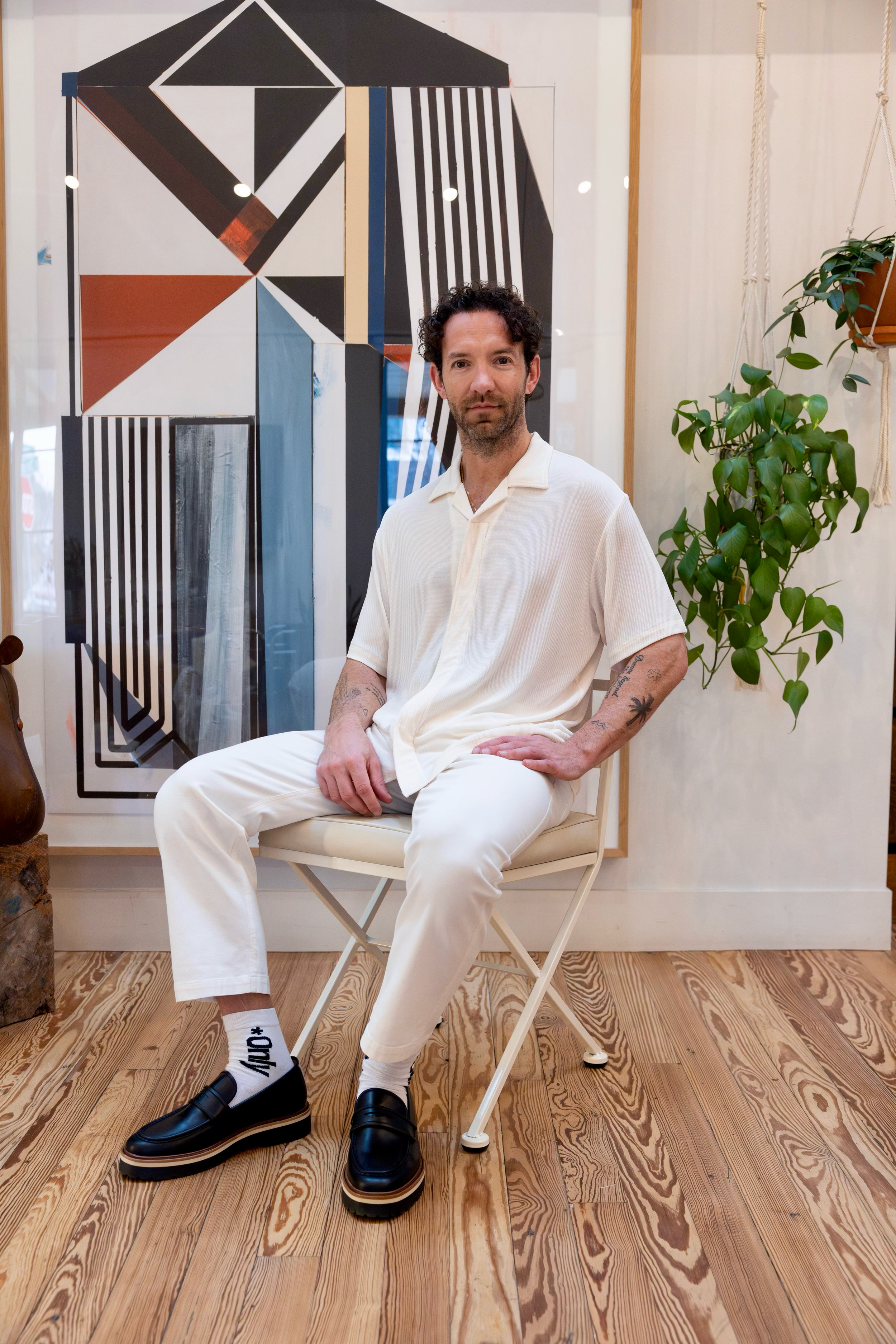 A man with tattoos on his arms and dark curly hair, dressed in a cream shirt and white pants, sitting on a cream chair in an indoor space. Behind him, there is a large abstract geometric painting and some green plants, including a pothos, with hardwood floors and light-colored walls.
