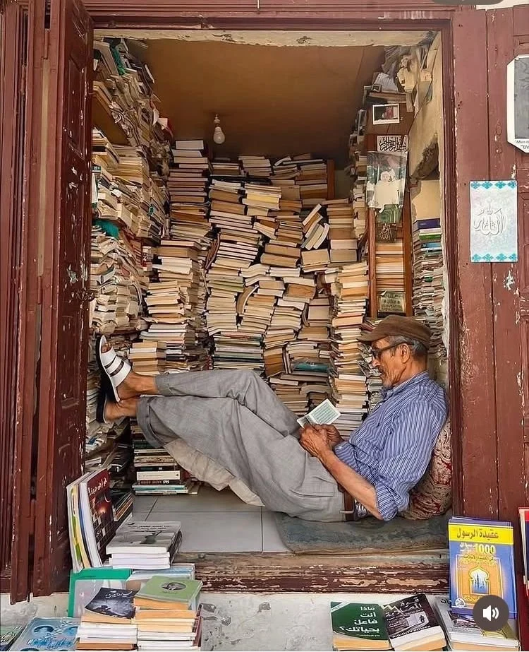 An elderly man reads a book while reclining in a small, cozy bookstore packed with stacks of books on shelves, benches, and the floor.