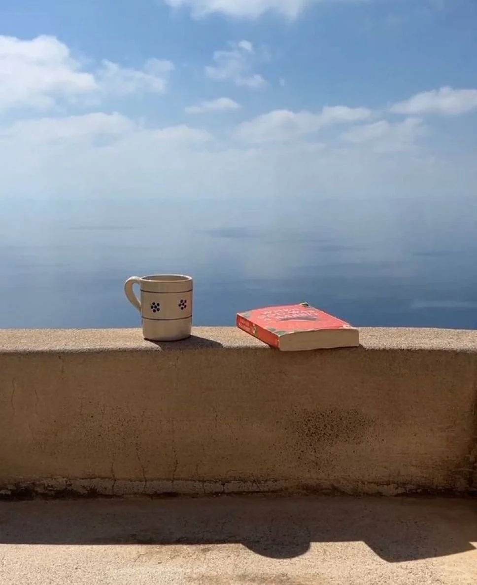 A coffee mug and a red book placed on a concrete barrier overlooking a calm sea with a partly cloudy sky.