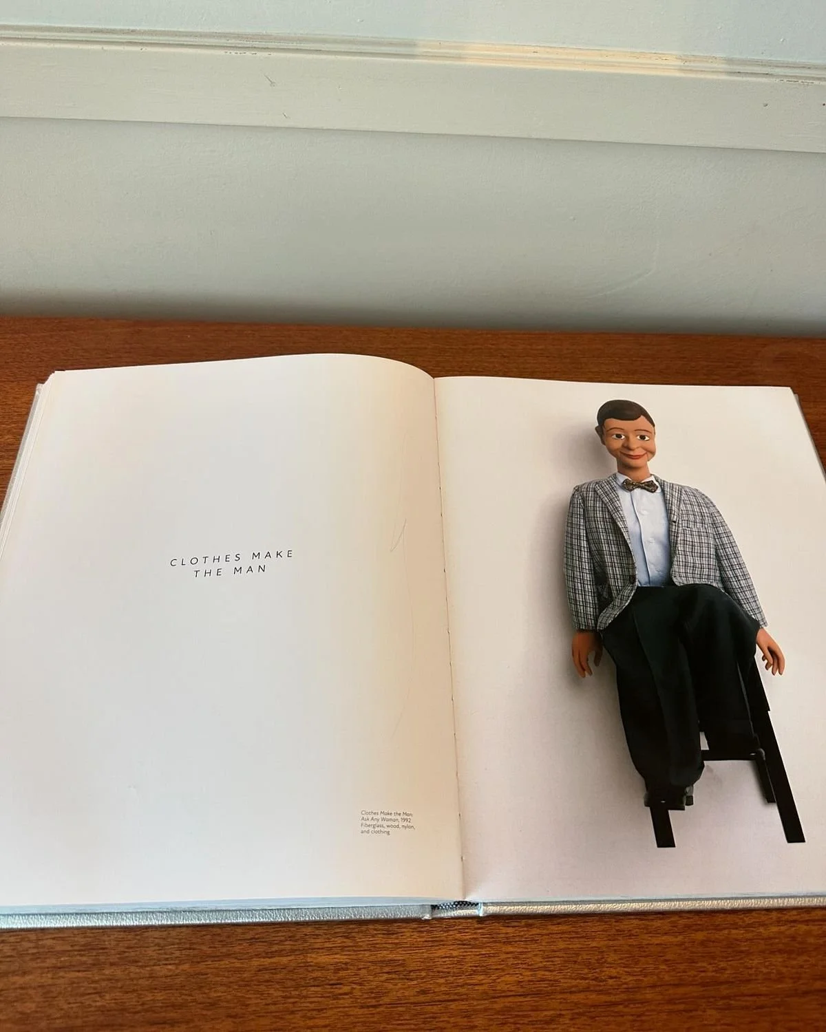 Open book with left page blank and right page showing a doll dressed in a plaid jacket, white shirt, bow tie, and black pants, sitting on a small chair. Text on the left page reads, "CLOTHES MAKE THE MAN".