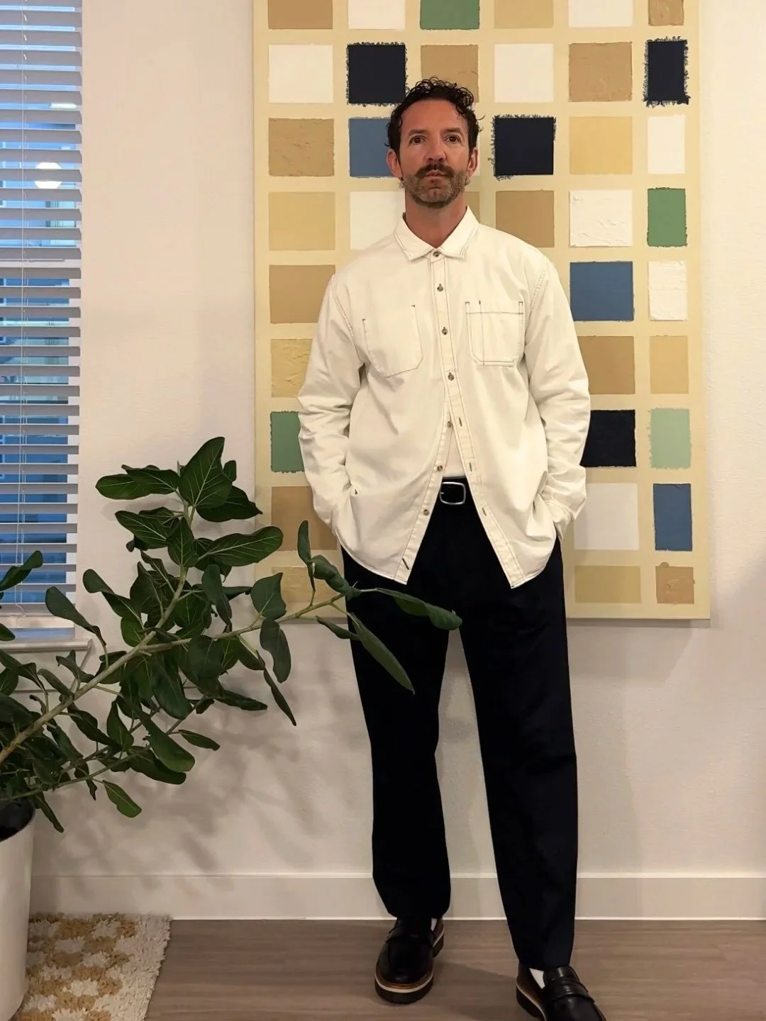 A man with dark curly hair and a beard stands with his hands in his pockets, wearing a white button-up shirt and black pants, in front of a colorful grid wall art, next to a green potted plant, in a well-lit room.