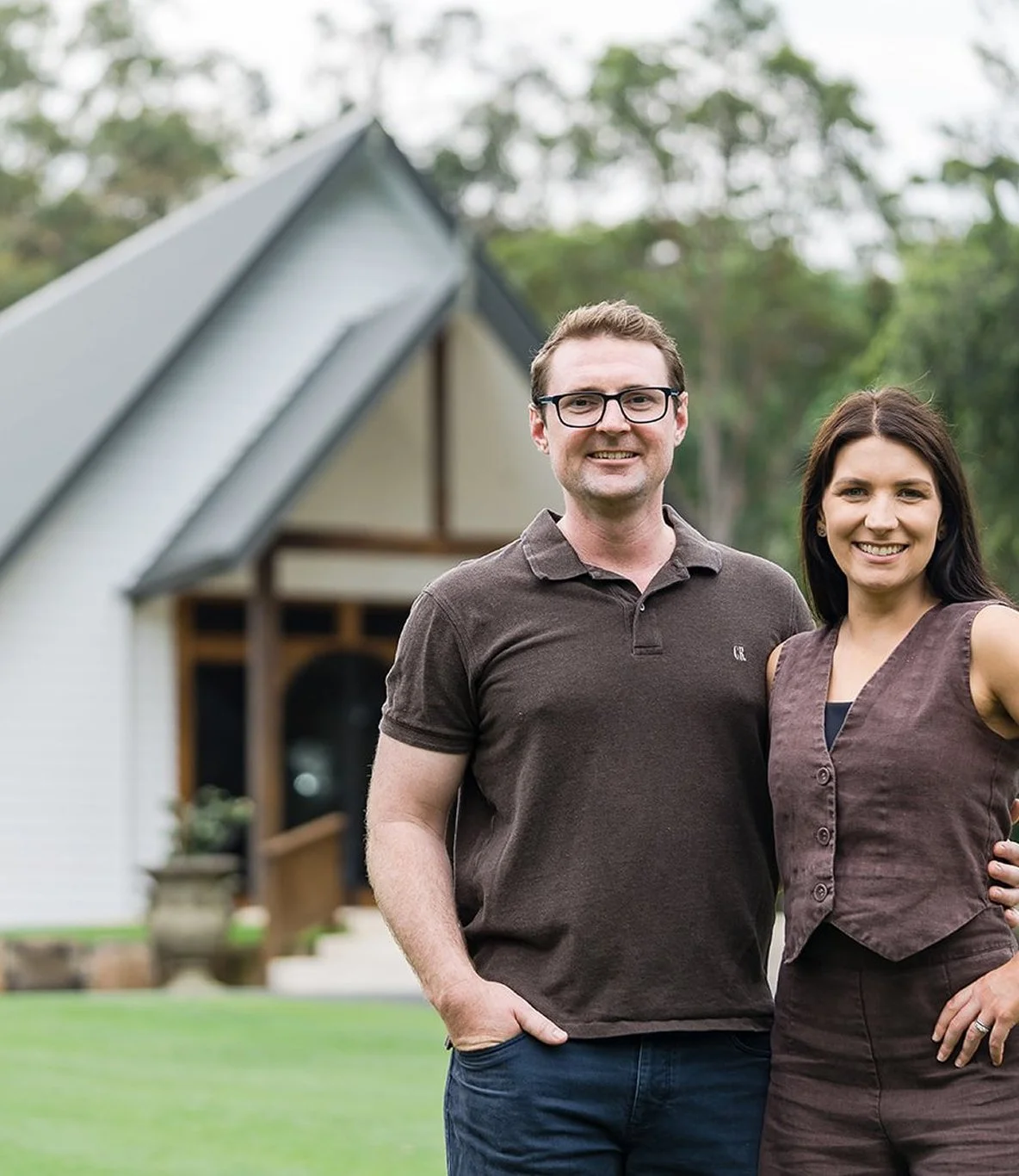 A smiling man wearing glasses and a dark polo shirt standing outdoors next to a smiling woman in a brown dress, with a small white house with a steep roof and trees in the background.