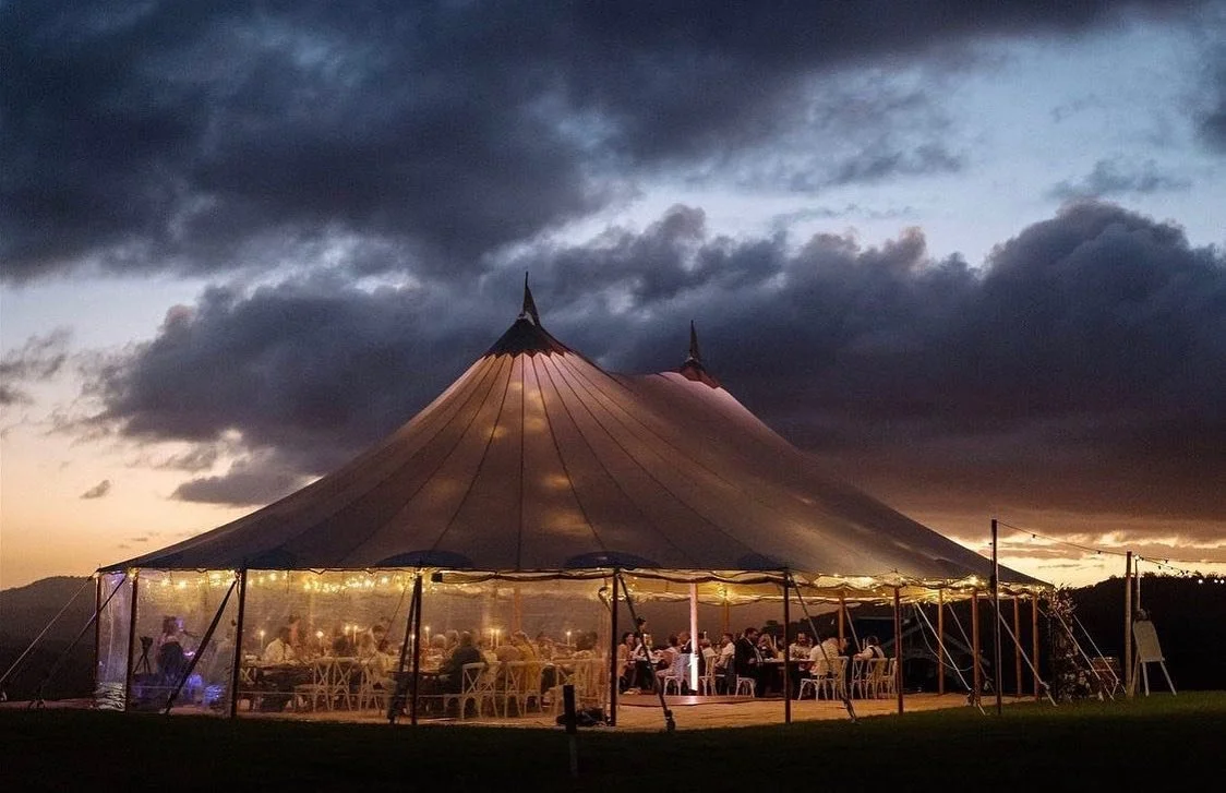 A large outdoor event tent illuminated at dusk, with people sitting inside and dark clouds overhead.