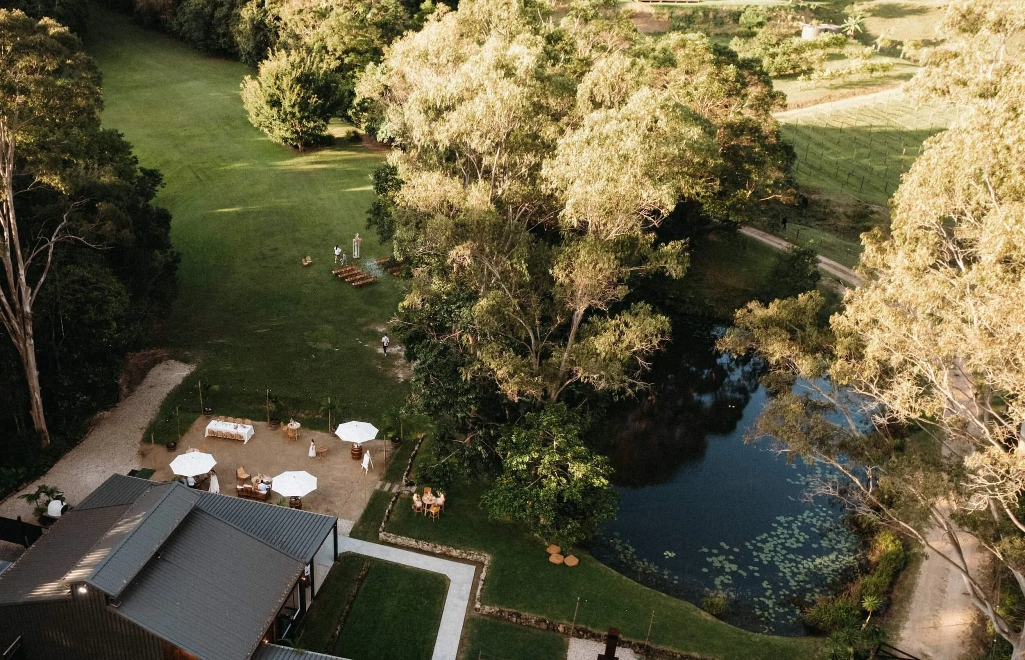 An aerial view of an outdoor event with white umbrellas, tables, and chairs set up near a dark pond, surrounded by trees and grassy areas.