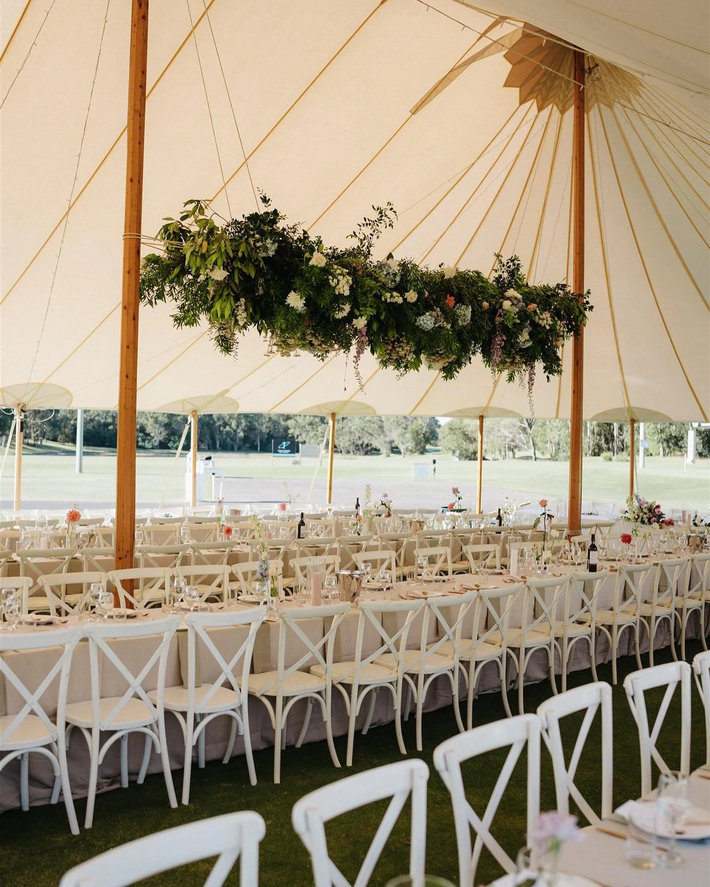 Wedding reception tent with long table set for guests, decorated with flowers and wine bottles, with a large hanging floral arrangement inside the tent.