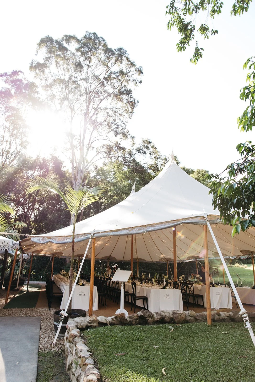 Outdoor event setup with a large white tent, arranged tables, and chairs, surrounded by trees on a sunny day.