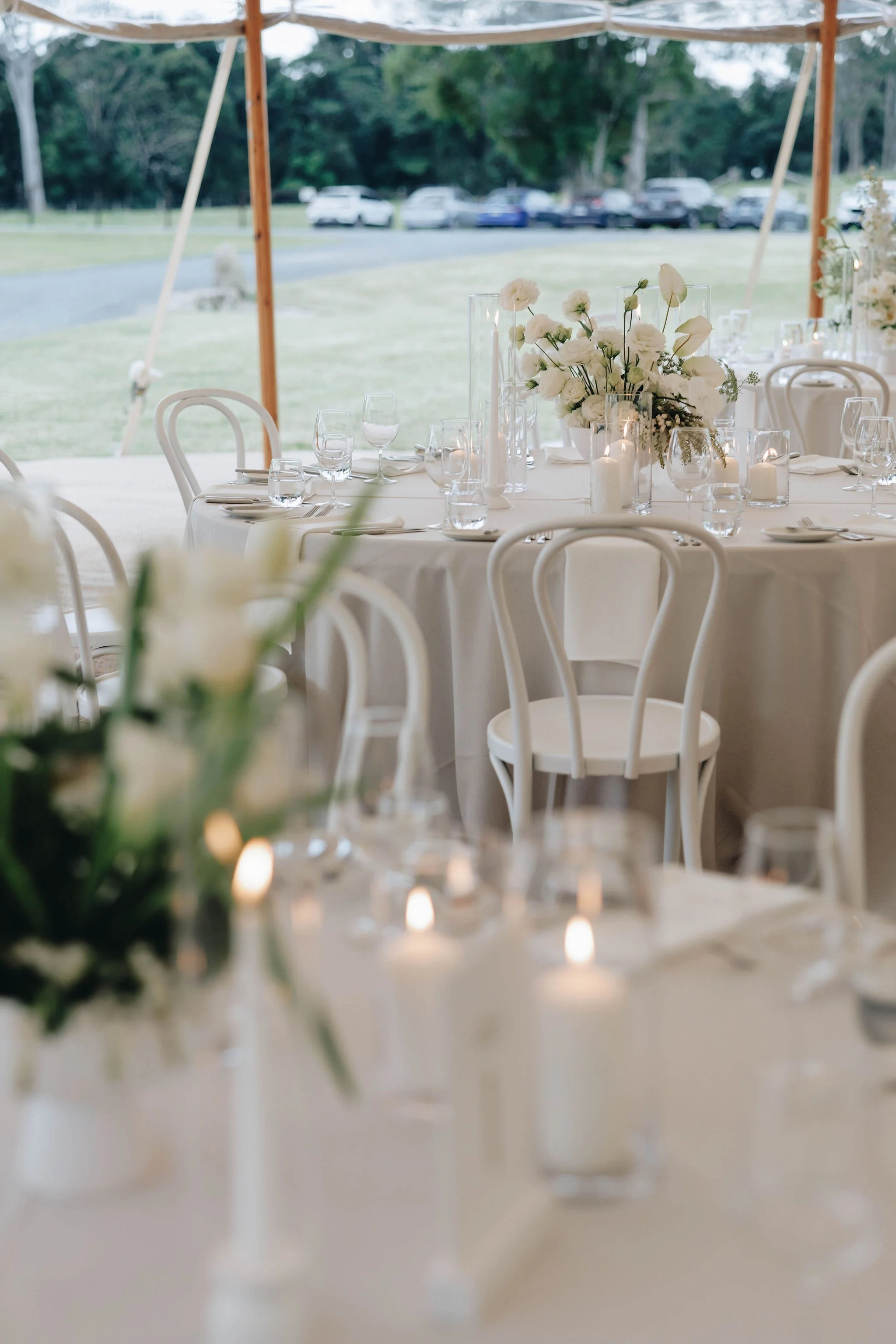 Elegant outdoor wedding reception setup with round tables, white tablecloths, floral centerpieces, candles, and white chairs under a canopy, with a grassy area, trees, and parked cars in the background.