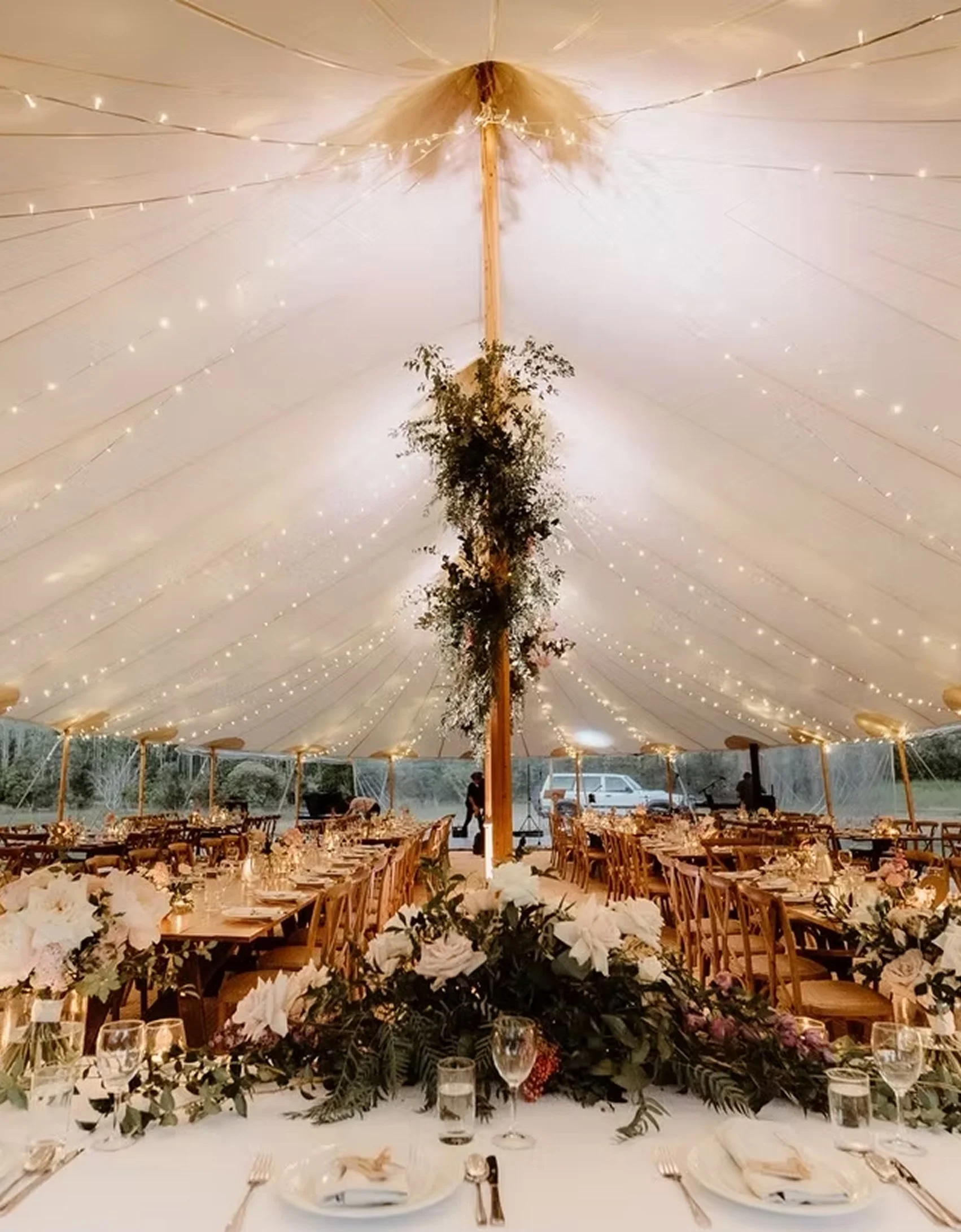 Wedding reception setup inside a large tent with string lights, wooden chairs, decorated tables, floral centerpieces, and a central pole decorated with greenery.