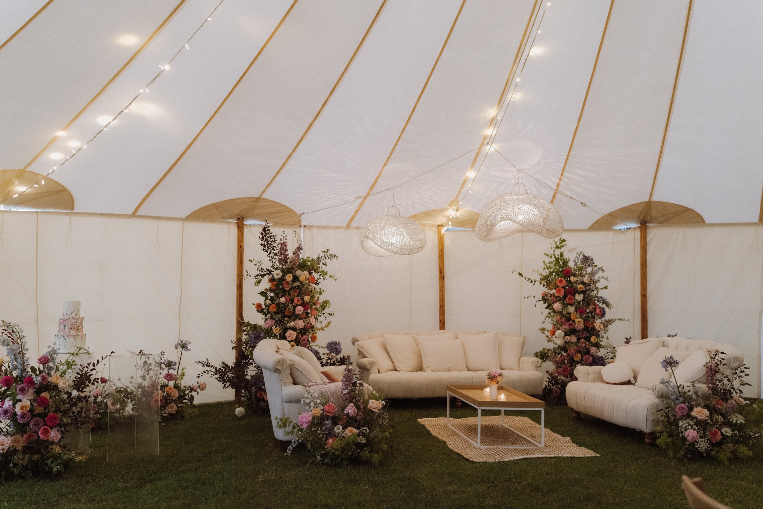 Decorated event tent interior with three cream-colored sofas, floral arrangements, a small table with candles, and hanging wicker lamps under string lights.