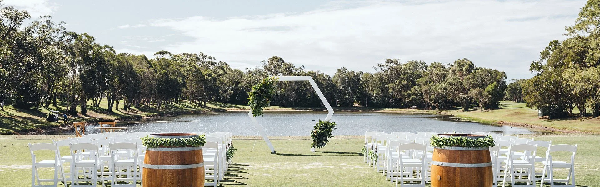 Outdoor wedding setup with white chairs arranged in front of a lake, decorated with greenery, with a geometric arch in the background on a sunny day.