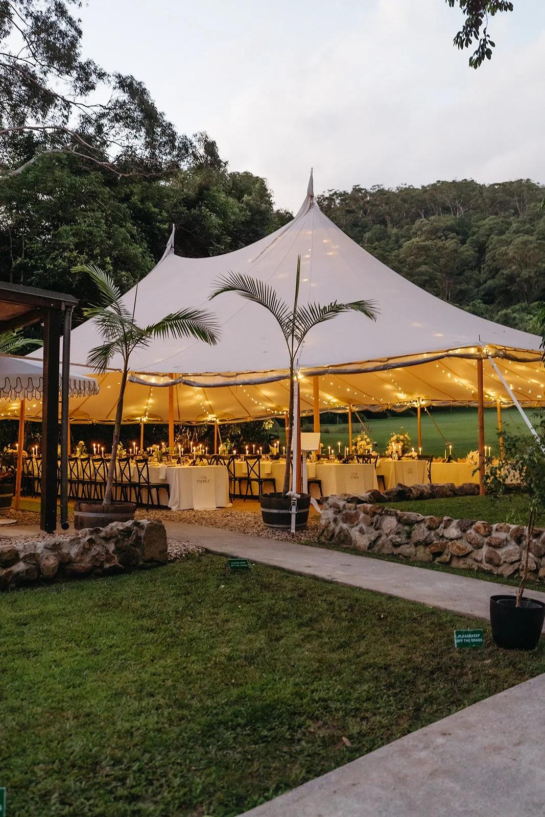 A large outdoor event tent decorated with string lights, set in a scenic area with trees and mountains in the background. Long tables with candles and floral centerpieces are arranged inside the tent for a celebration or gathering.
