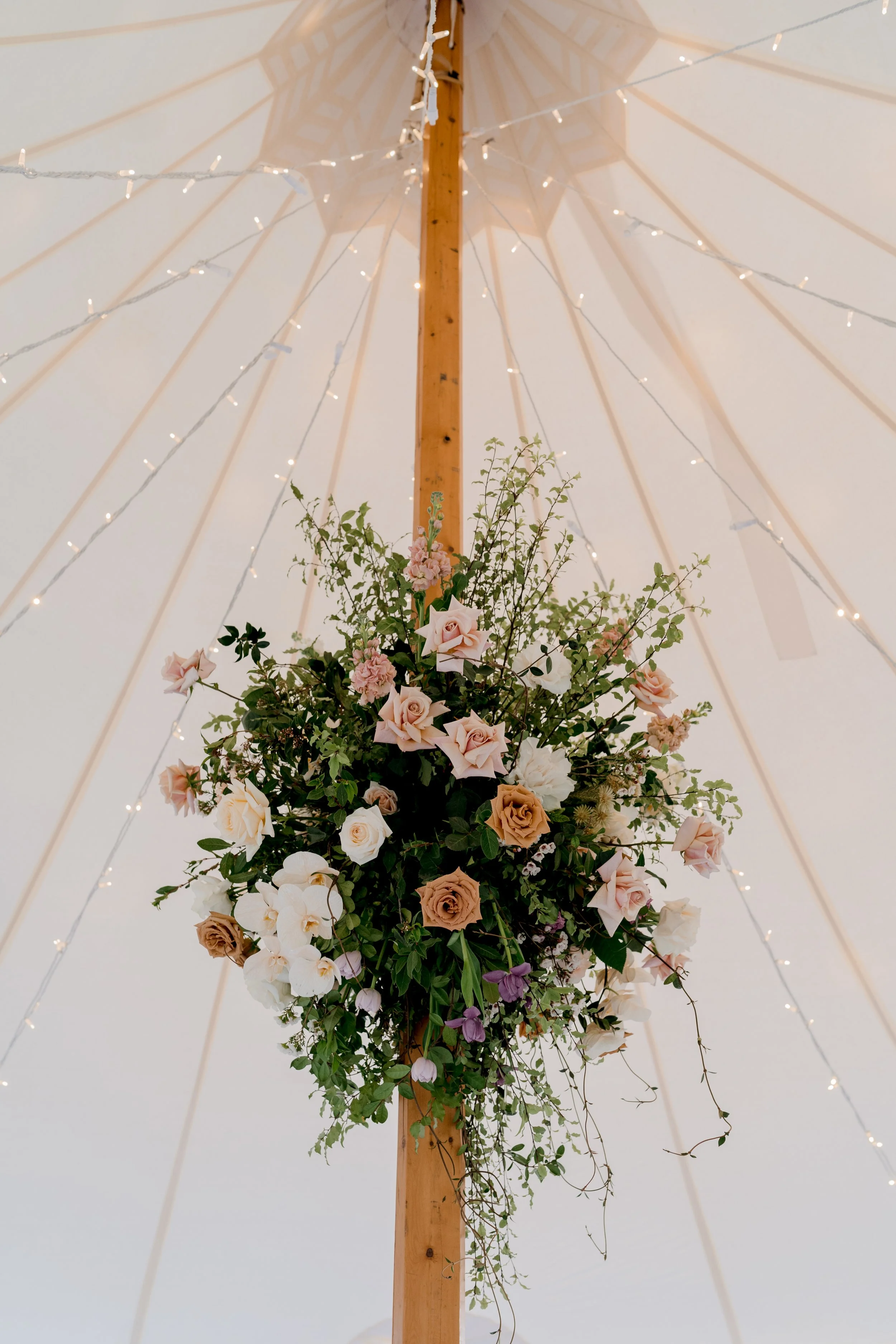Hanging floral arrangement with pink, white, and peach roses and greenery under a cream-colored tent with string lights.