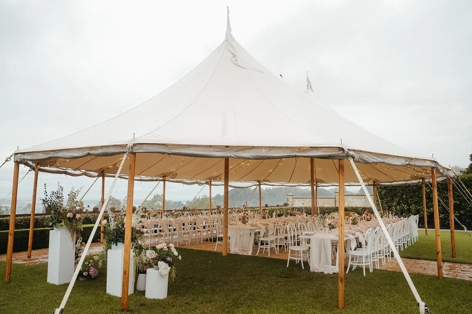 A large white event tent set up outdoors on grass, decorated with floral arrangements and string lights, with empty tables and chairs arranged inside.