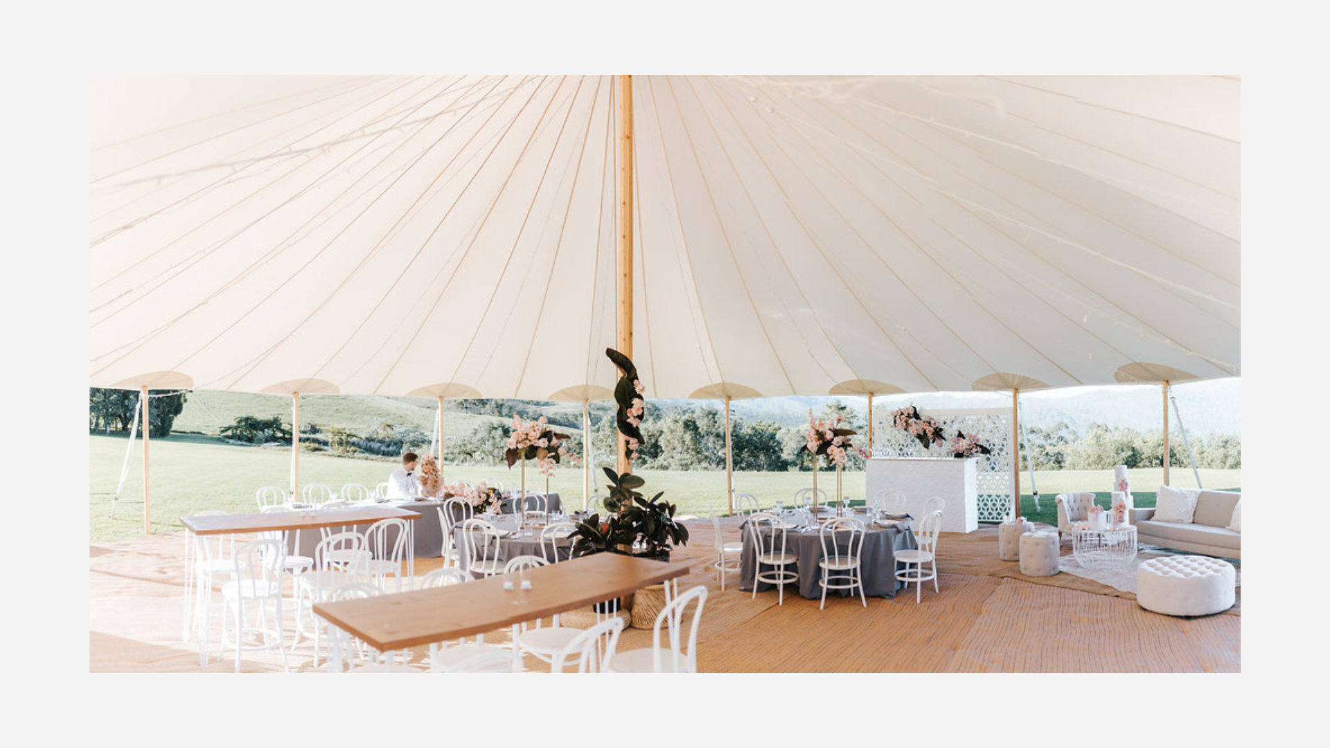 Elegant outdoor event setup under a large white tent with round tables decorated with pink flowers, surrounded by white chairs, and cozy lounge seating with cushions and ottomans, overlooking a green landscape.