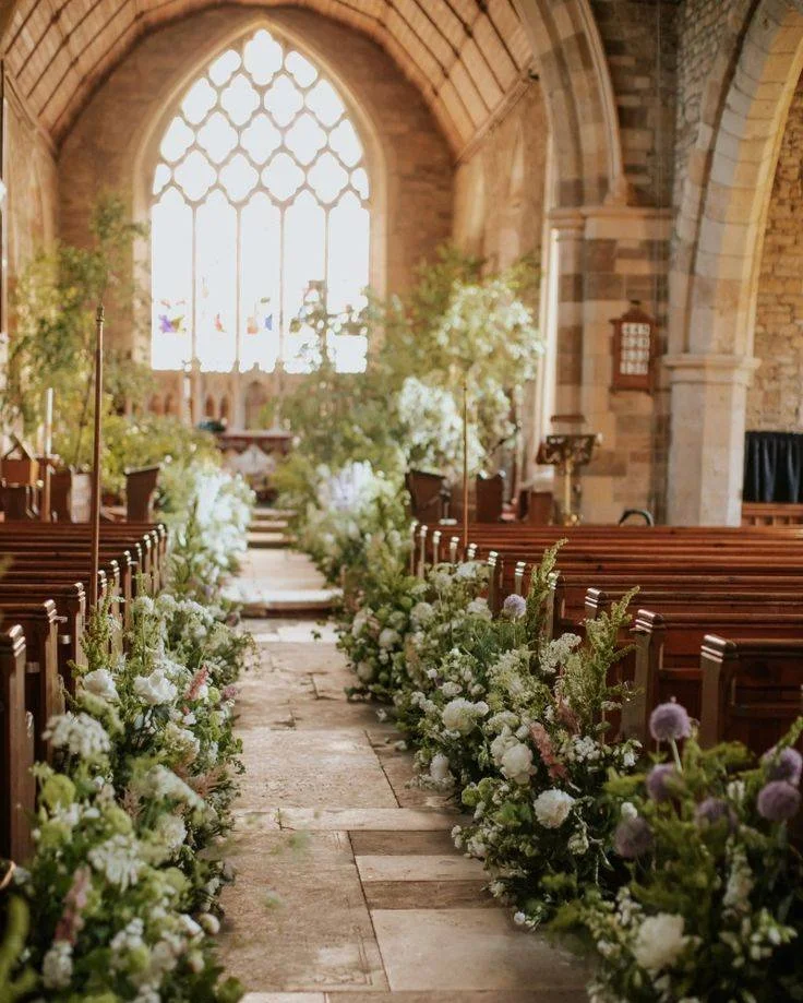 Interior of a church decorated for a wedding, with a flower-strewn aisle leading to the altar and large stained glass window in the background.